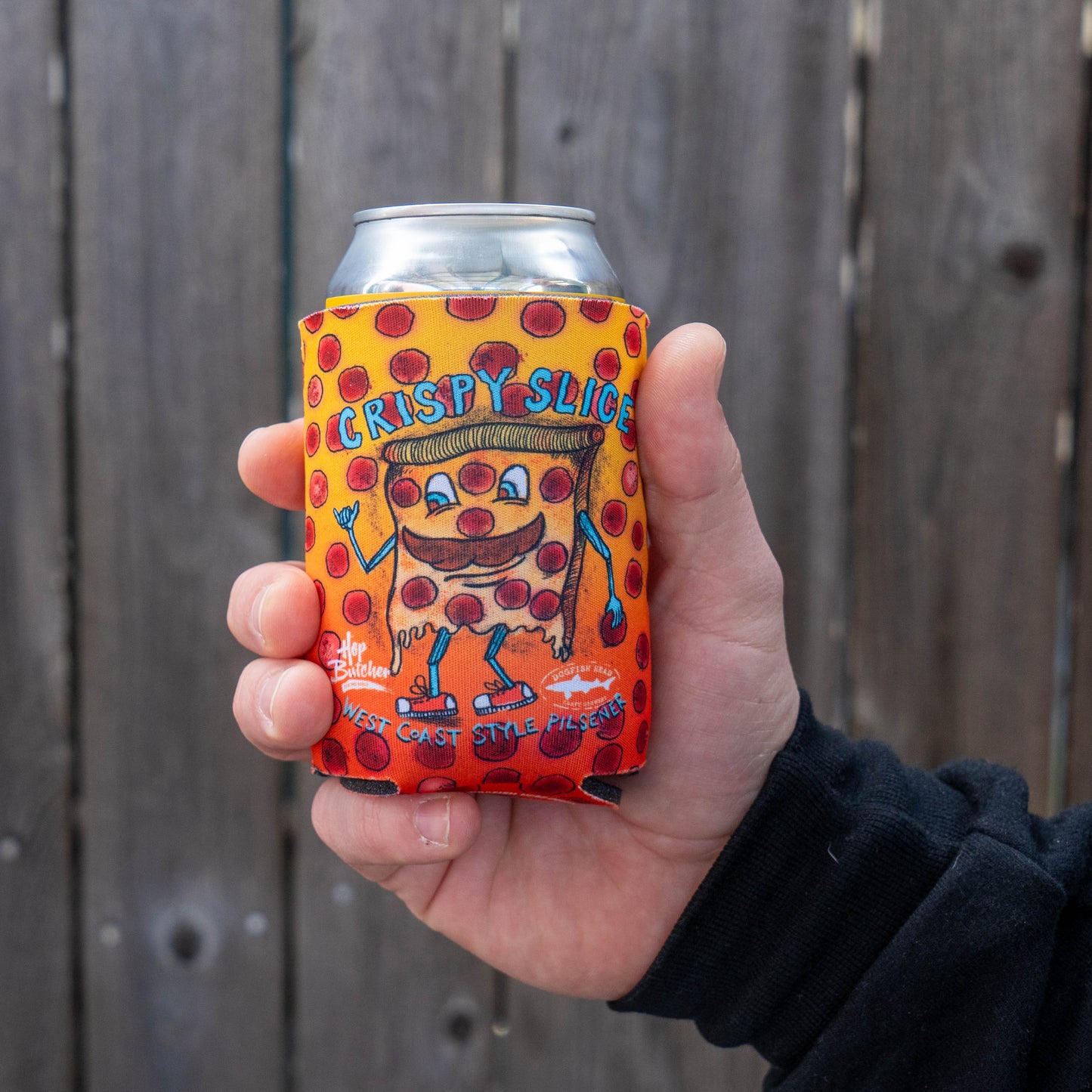 A hand holds a Grandstand Glassware & Apparel Crispy Slice 12oz Can Cooler, featuring playful lettering and polka dots with a cartoon pizza slice, wrapped around a can against a wooden fence—perfect for keeping drinks cold.