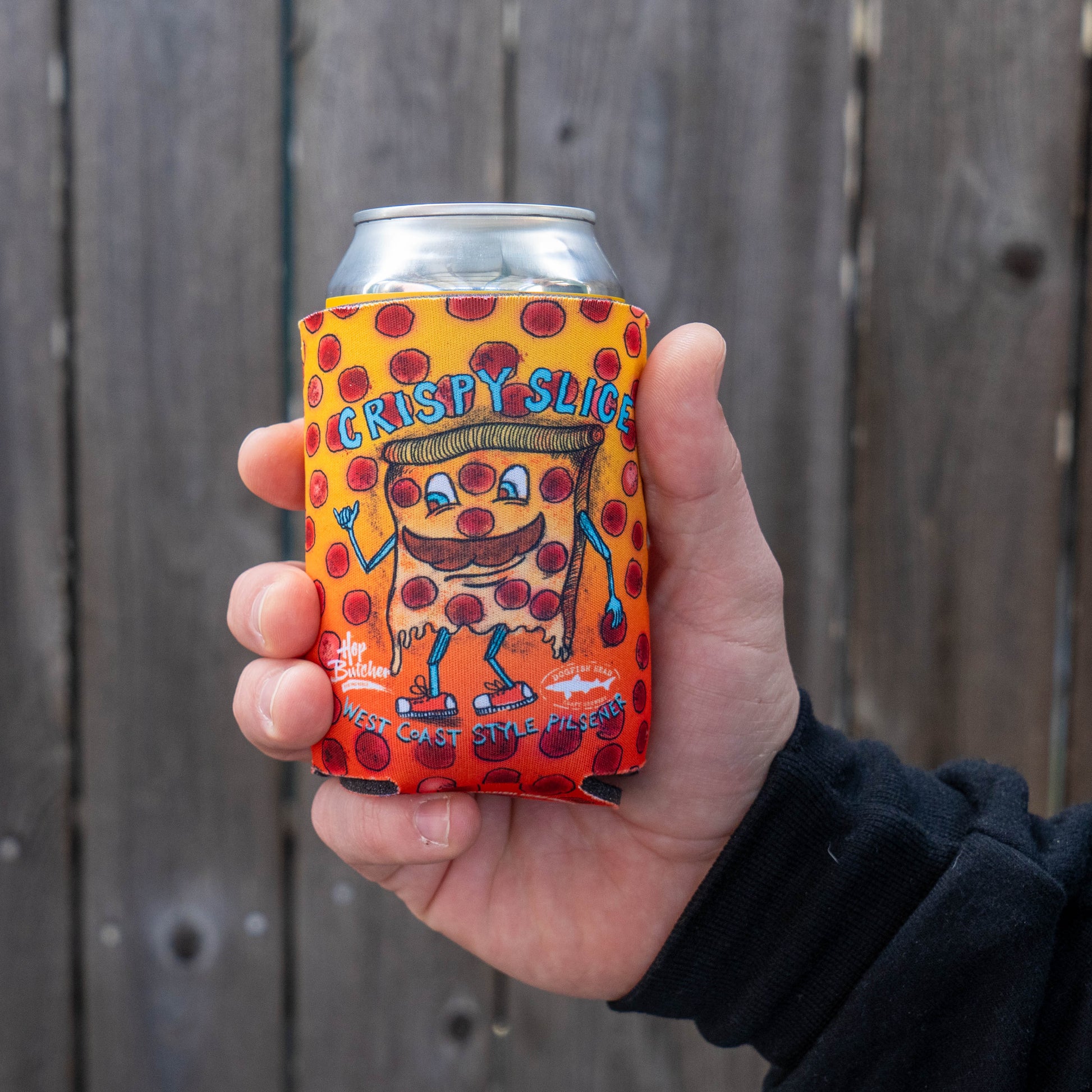 A hand holds a Grandstand Glassware & Apparel Crispy Slice 12oz Can Cooler, featuring playful lettering and polka dots with a cartoon pizza slice, wrapped around a can against a wooden fence—perfect for keeping drinks cold.