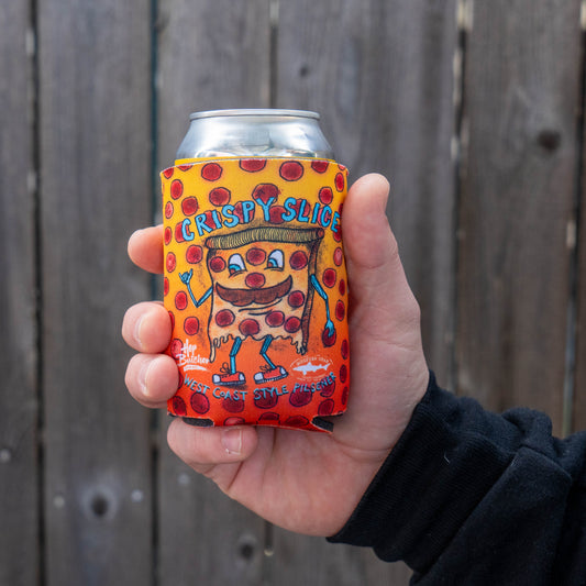 A hand holds a Grandstand Glassware & Apparel Crispy Slice 12oz Can Cooler, featuring playful lettering and polka dots with a cartoon pizza slice, wrapped around a can against a wooden fence—perfect for keeping drinks cold.