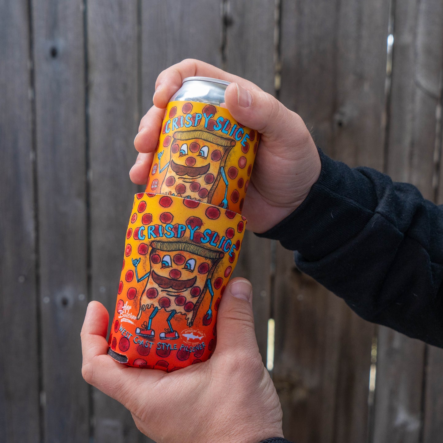 A person holds a 12oz can with cartoon pizza graphics and the text "Crispy Slice," wrapped in a Grandstand Glassware & Apparel Crispy Slice 12oz Can Cooler, against a wooden fence background.