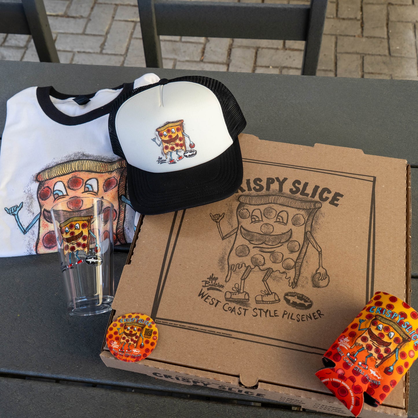 The Crispy Slice Merch Bundle by GO WEST T-SHIRT COMPANY includes a pizza box, hat, T-shirt, pint glass, stickers, and can holder—each featuring the signature cartoon pizza slice character.