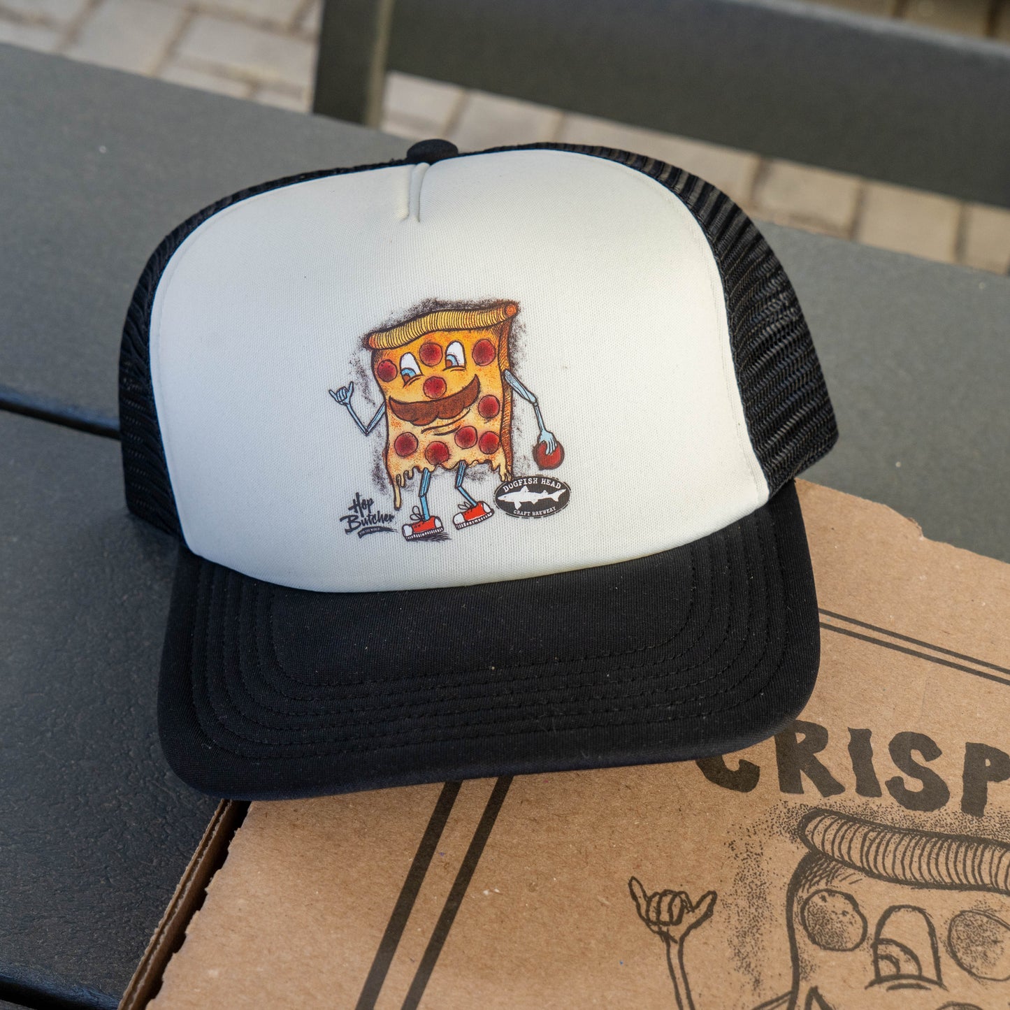 Dogfish Head x Crispy Pizza trucker hat with cartoon pizza slice graphic, white foam front panel and black brim on a Crispy Pizza box