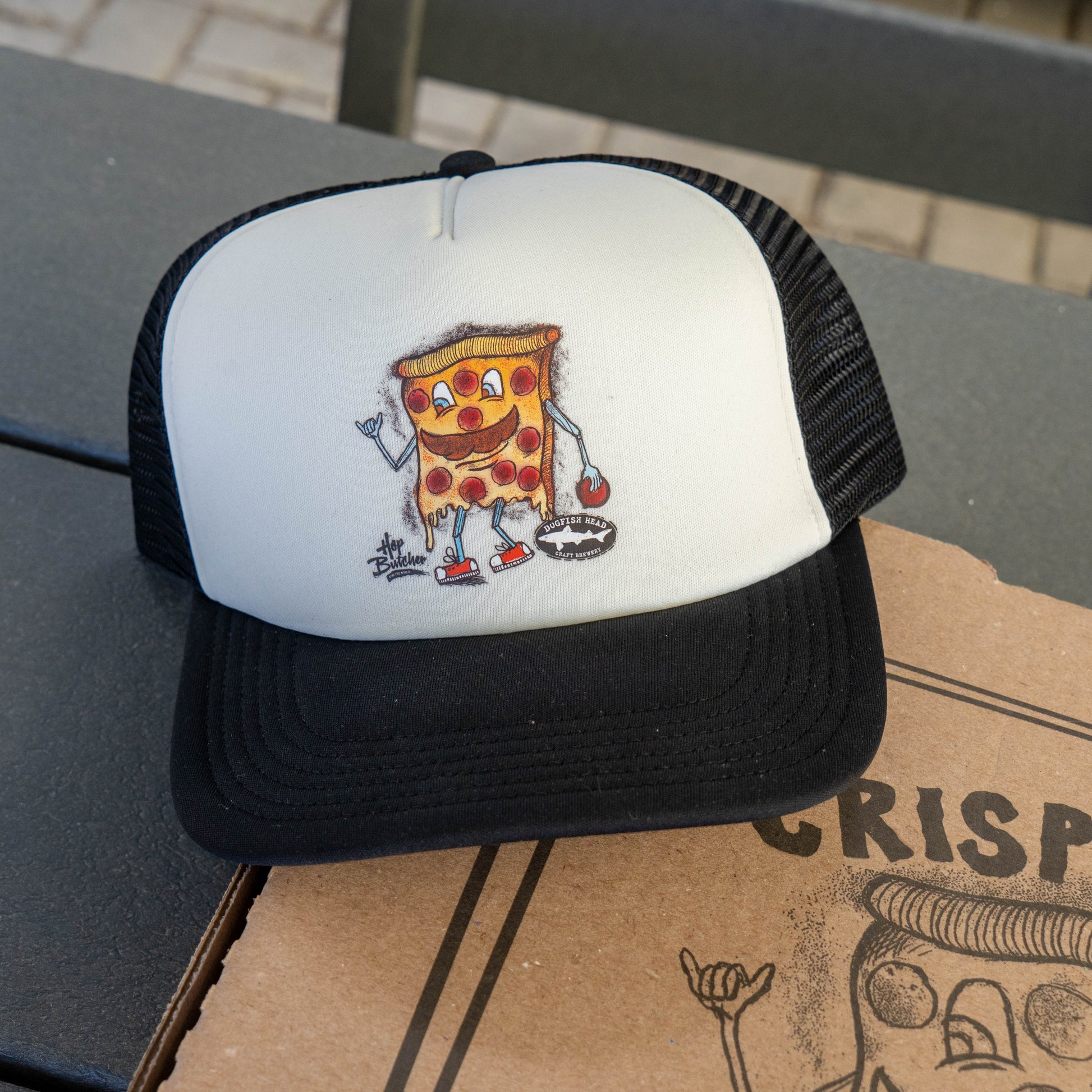 Dogfish Head x Crispy Pizza trucker hat with cartoon pizza slice graphic, white foam front panel and black brim on a Crispy Pizza box