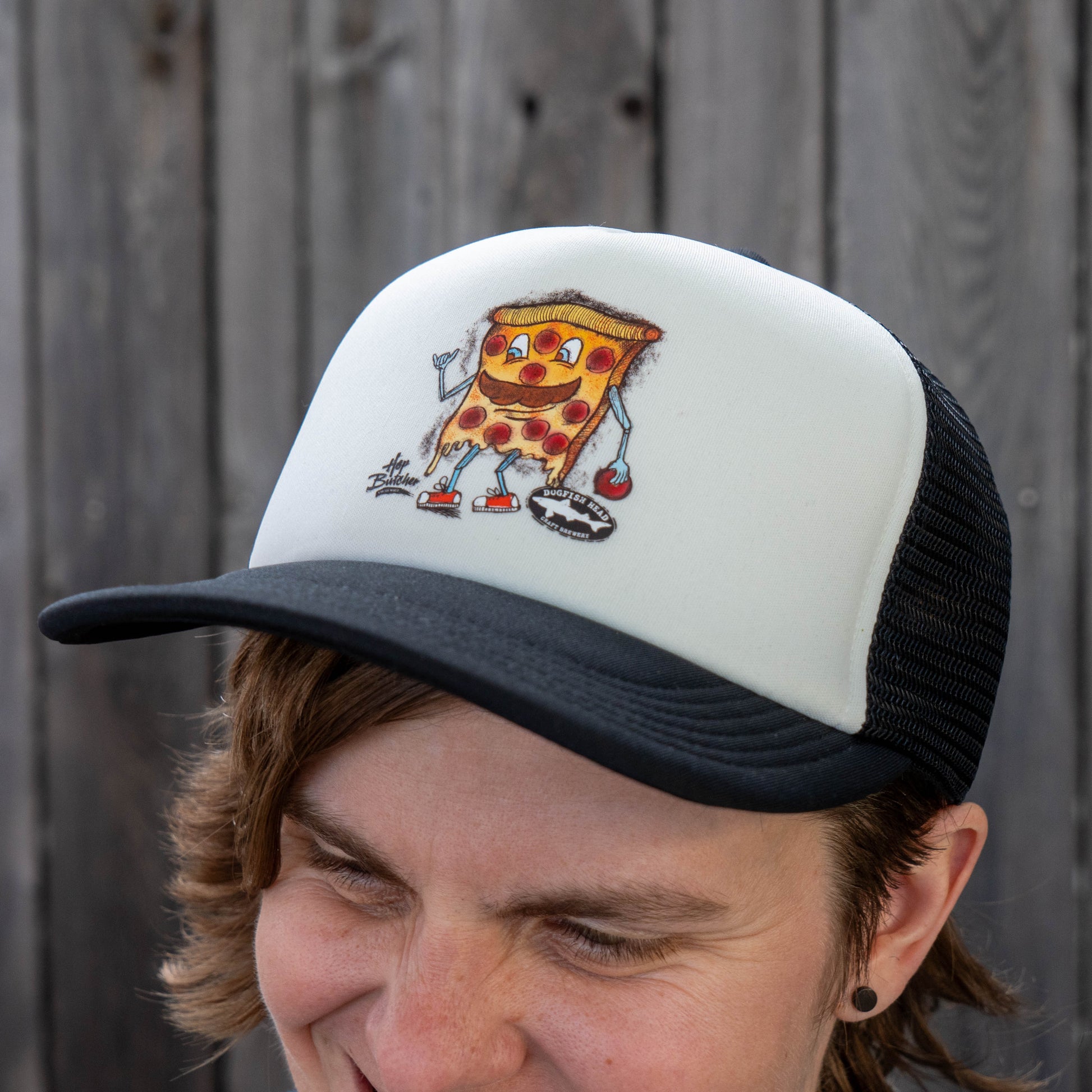Dogfish Head x Crispy Pizza trucker hat with cartoon pizza slice graphic, white foam front panel and black brim, worn outdoors close-up