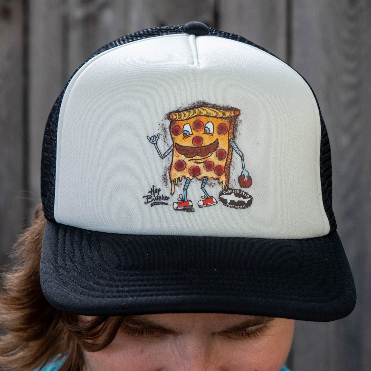 Dogfish Head x Crispy Pizza trucker hat with cartoon pizza slice graphic, white foam front panel and black brim, worn outdoors close-up