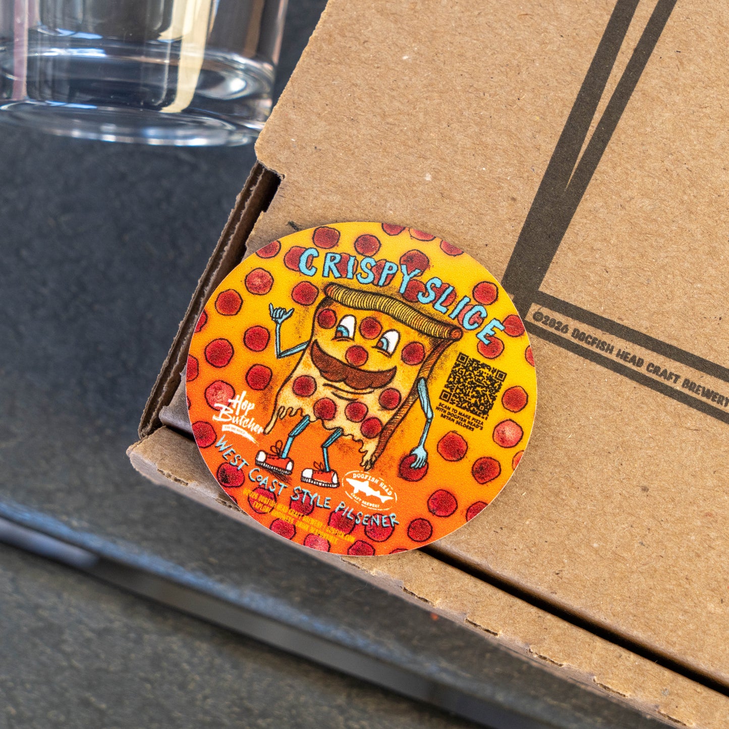 Dogfish Head x Crispy Pizza magnet with cartoon pepperoni pizza slice character on orange background sitting on pizza box next to glass