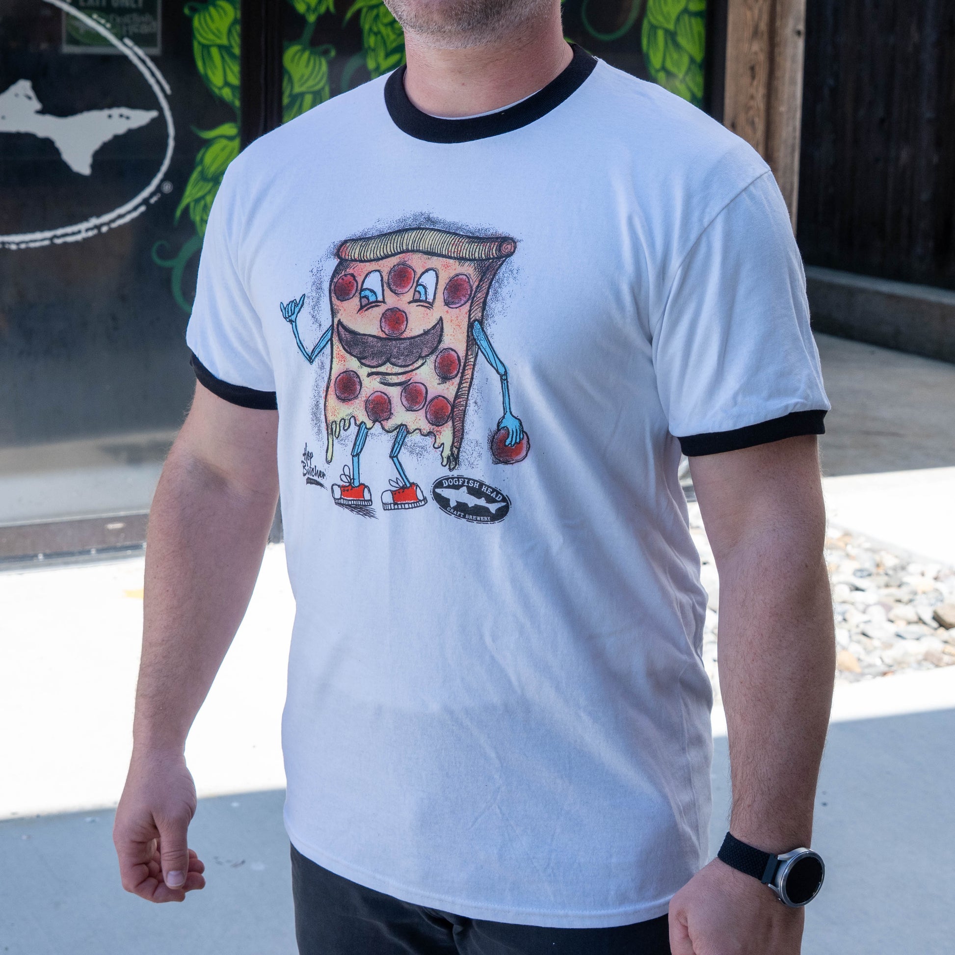 A person wears the GO WEST T-SHIRT COMPANY Crispy Slice Tee Shirt, a white tee with black trim featuring a cartoon pizza slice with arms, legs, and a face giving a thumbs up, standing by a bowl outdoors near a building.