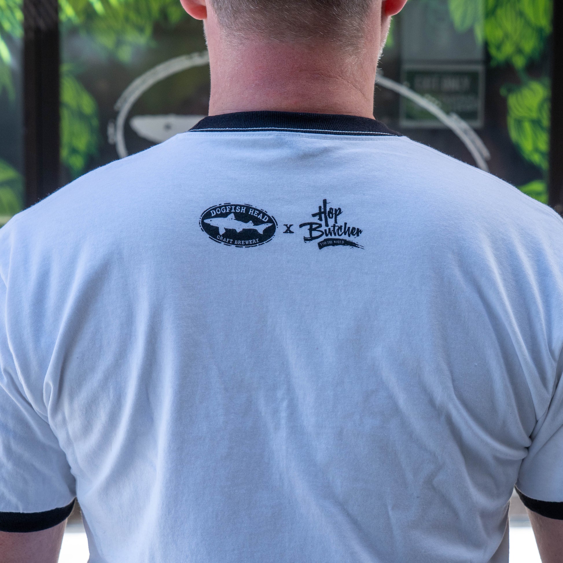A person wearing a white Crispy Slice Tee Shirt by GO WEST T-SHIRT COMPANY stands with their back to the camera. The shirt has logos and collaboration text near the neck, with green leaves and a partial logo blurred in the background.