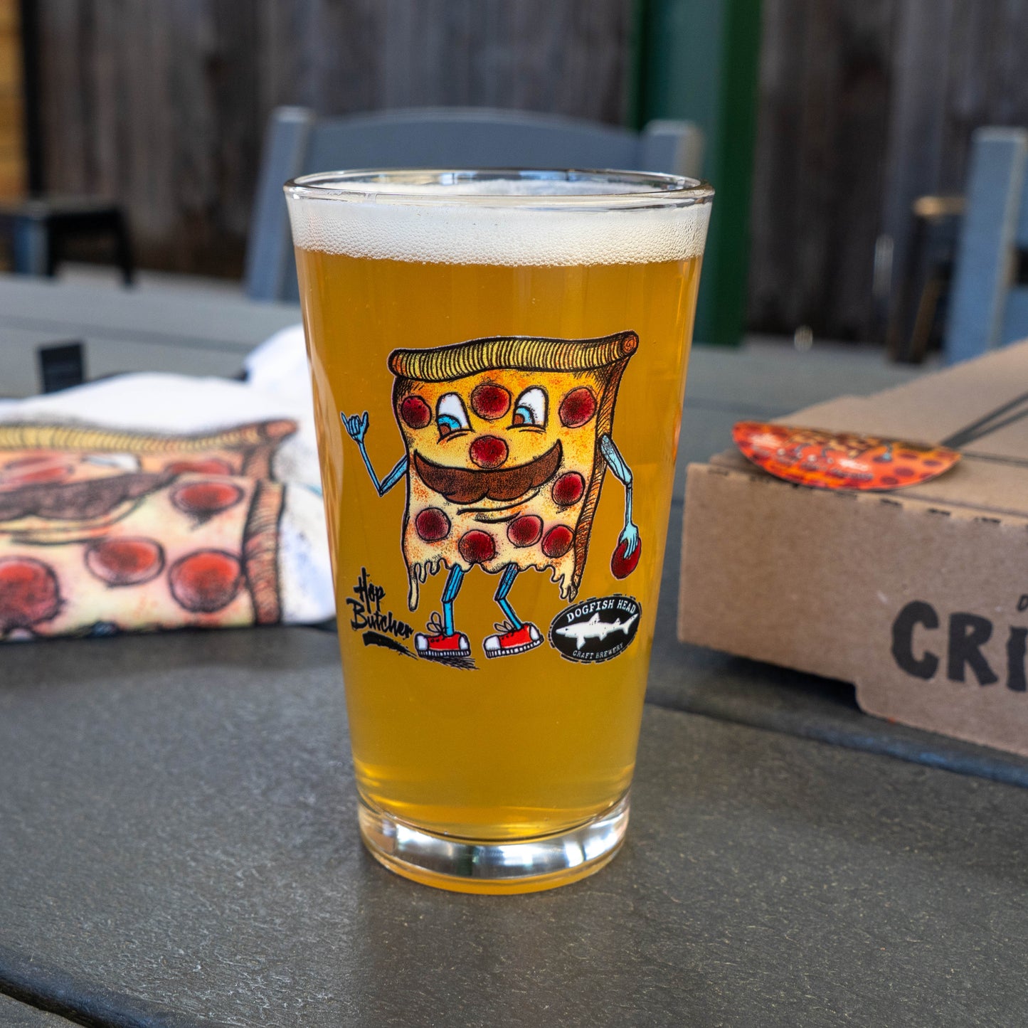 A pint glass of beer rests outdoors on a table with Crispy Slice, a cartoon pizza character, from the GO WEST T-SHIRT COMPANY’s Crispy Slice Merch Bundle. A pizza box and a pizza-patterned cloth are also nearby.
