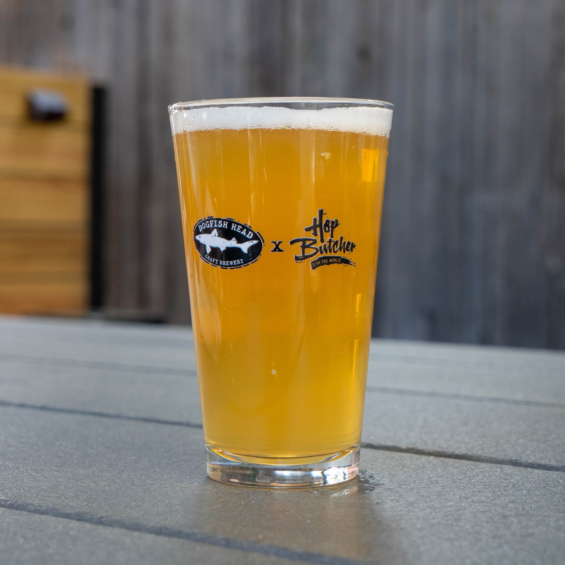 A hazy golden beer in a pint glass sits on a gray table. The glass features logos for GO WEST T-SHIRT COMPANY and Crispy Slice Merch Bundle. Blurred wood paneling is visible in the background.