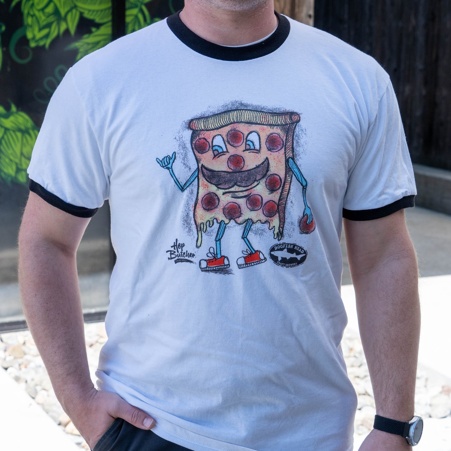 A person wears the GO WEST T-SHIRT COMPANY Crispy Slice Merch Bundle: a white ringer tee featuring a smiling cartoon pepperoni pizza with arms, legs, sneakers, waving and holding a red drink while outdoors.