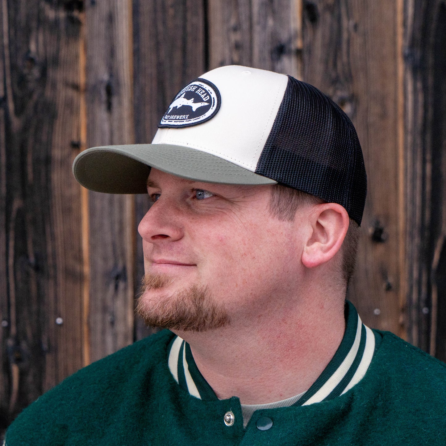 Side profile of a man wearing a black and white Dogfish Head trucker hat with an olive colored rim. The mesh on the back half is black and the front is solid white with a black dogfish head craft brewery logo.