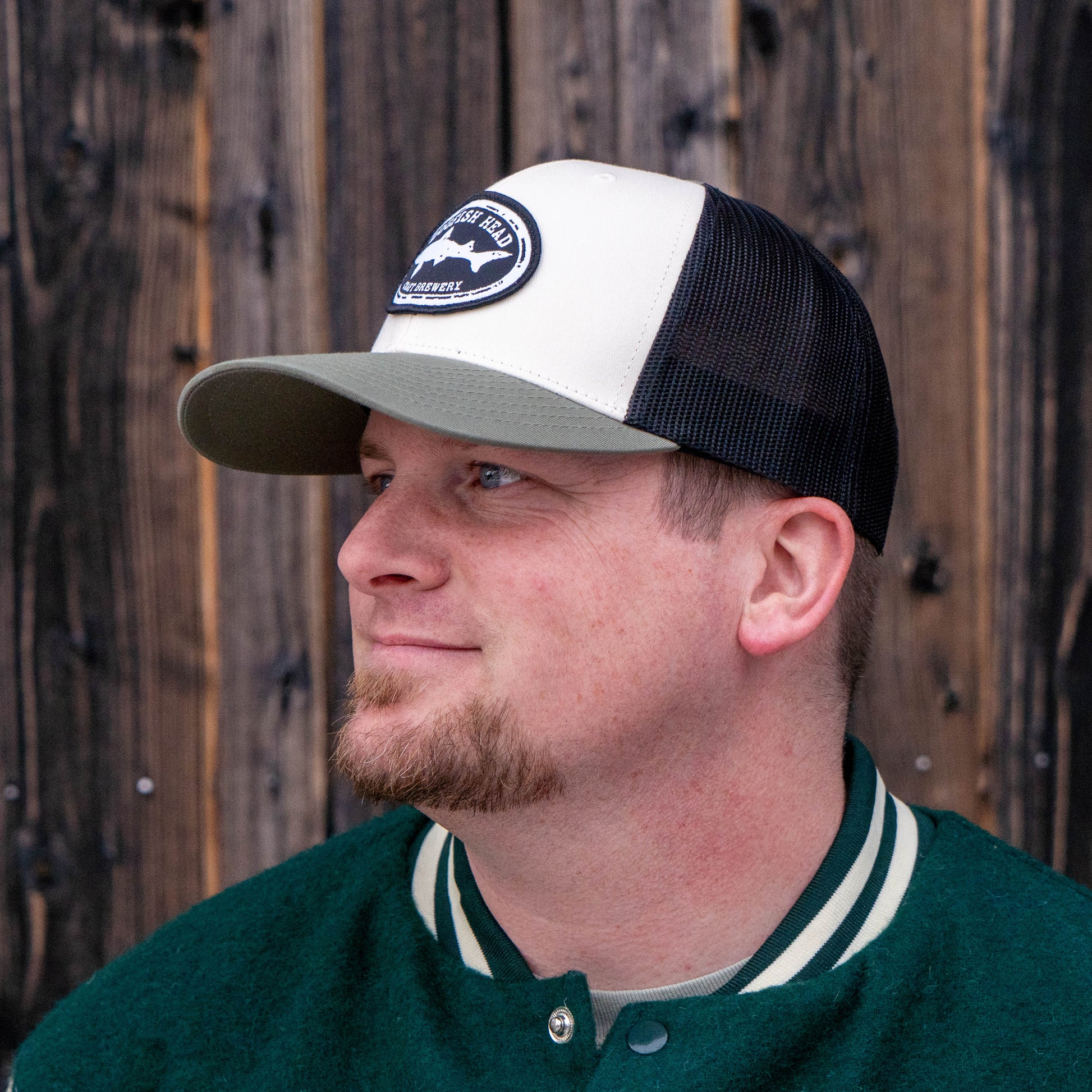 Side profile of a man wearing a black and white Dogfish Head trucker hat with an olive colored rim. The mesh on the back half is black and the front is solid white with a black dogfish head craft brewery logo.