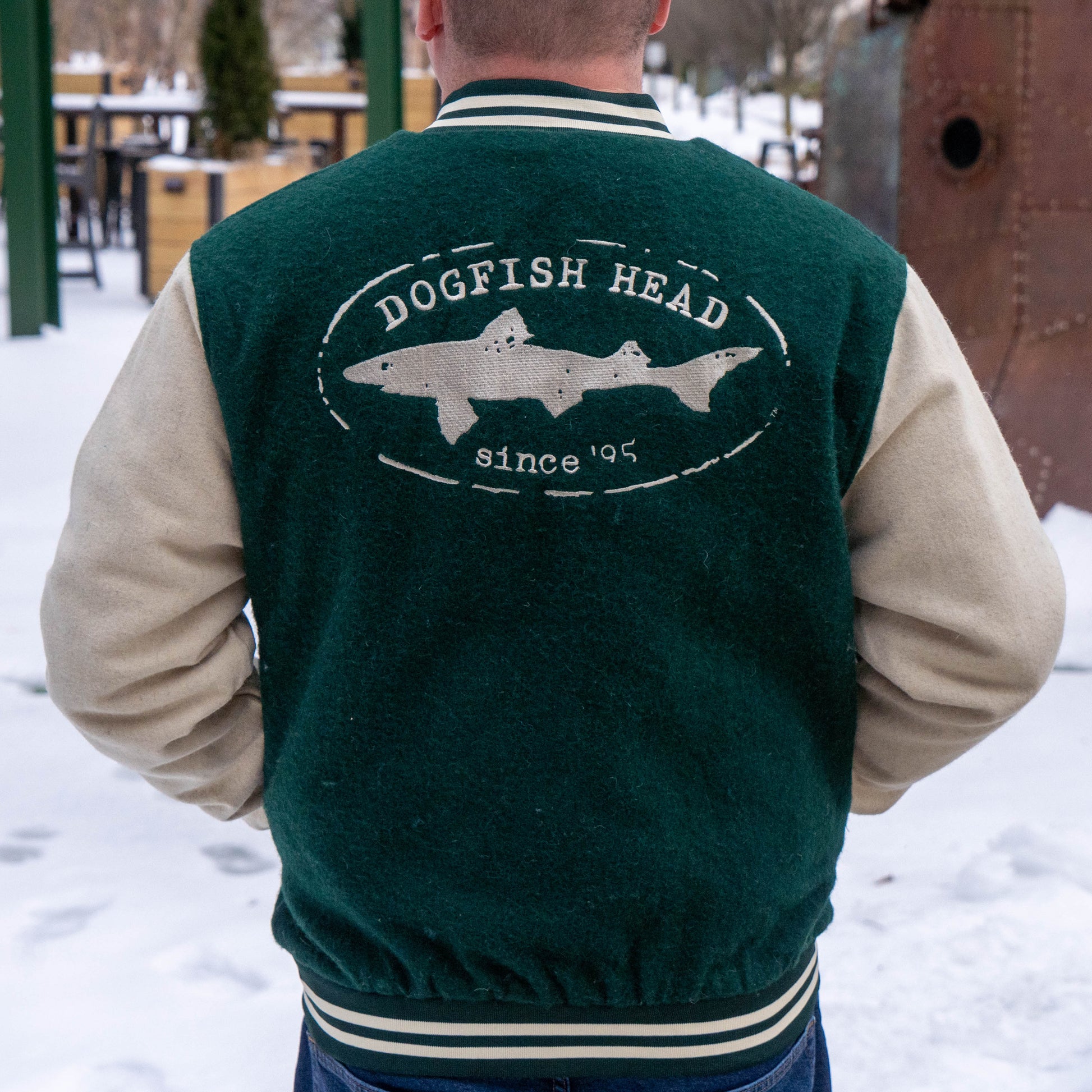 A person stands outside in the snow wearing Bottoms Up's Varsity Jacket 2 in green and cream, featuring "Dogfish Head since 95" and a shark image on the back.