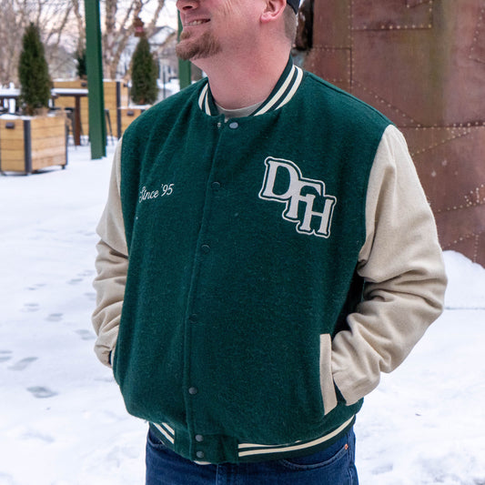 A man stands outside in the snow, smiling with his hands in his pockets, wearing the green and beige Varsity Jacket 2 by Bottoms Up.