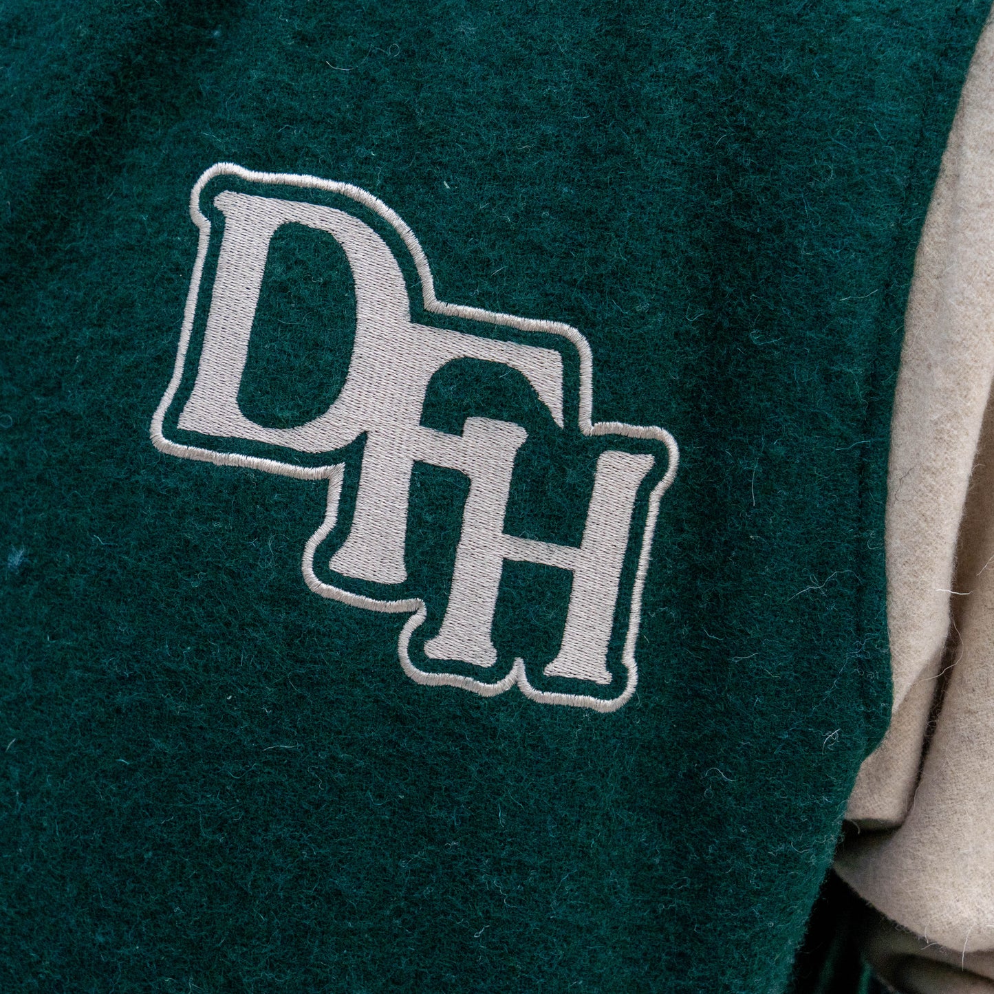 Close-up of a green fabric patch with large white outlined "DFH" letters sewn onto the Bottoms Up Varsity Jacket 2, with beige fabric from the jacket visible at the side.