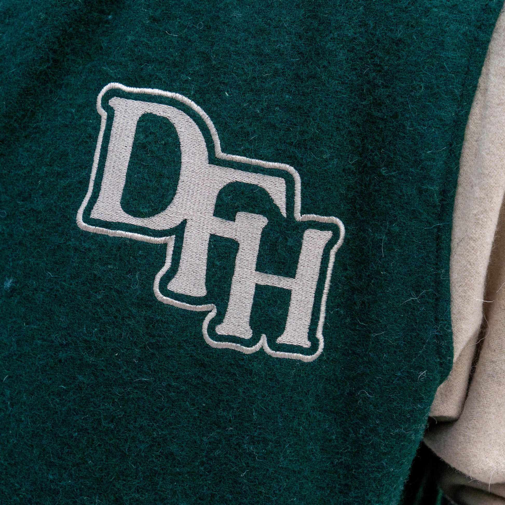 Close-up of a green fabric patch with large white outlined "DFH" letters sewn onto the Bottoms Up Varsity Jacket 2, with beige fabric from the jacket visible at the side.