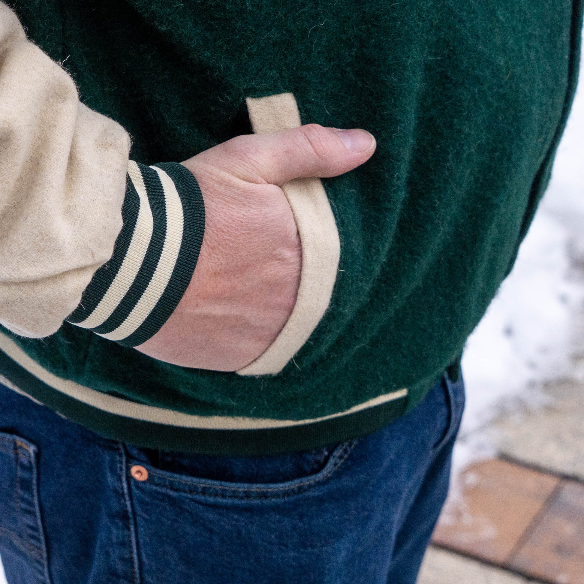 Someone stands outdoors with one hand in their pocket, wearing blue jeans and the Bottoms Up Varsity Jacket 2 in green and beige. Snow and pavement appear in the background.