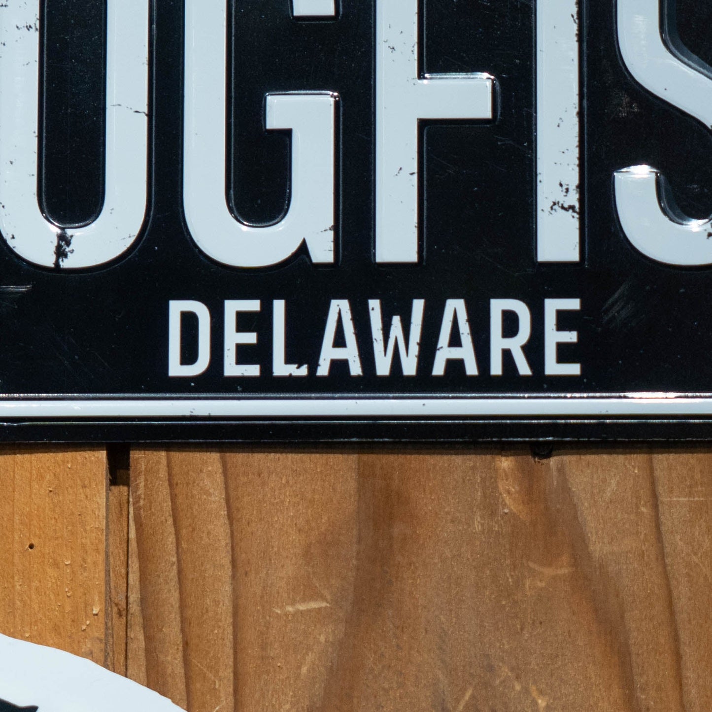 A close-up of the Merchtank Black DOGFISH License Plate Tacker shows "DELAWARE" in white on a black background, partially visible and mounted on wood—ideal decor for Dogfish or Delaware beer fans, or to pair with a metal wall tacker.