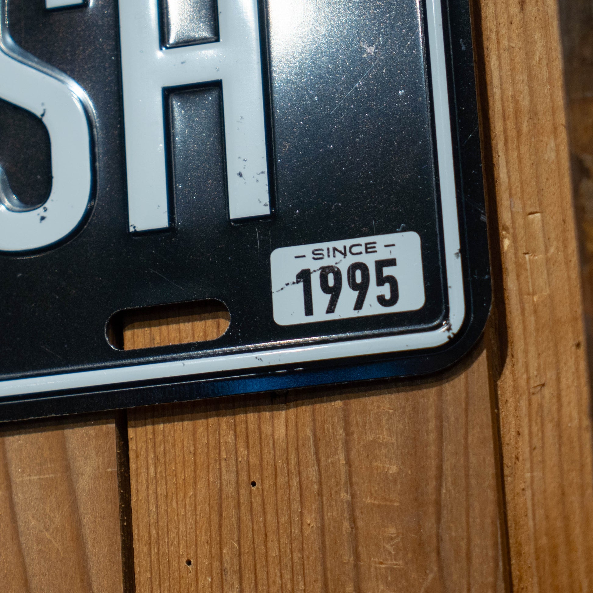 Close-up of a Black DOGFISH License Plate Tacker by Merchtank mounted on wood, featuring 'SH' and a white corner label reading 'SINCE 1995,' subtly referencing Dogfish’s Delaware beer heritage.