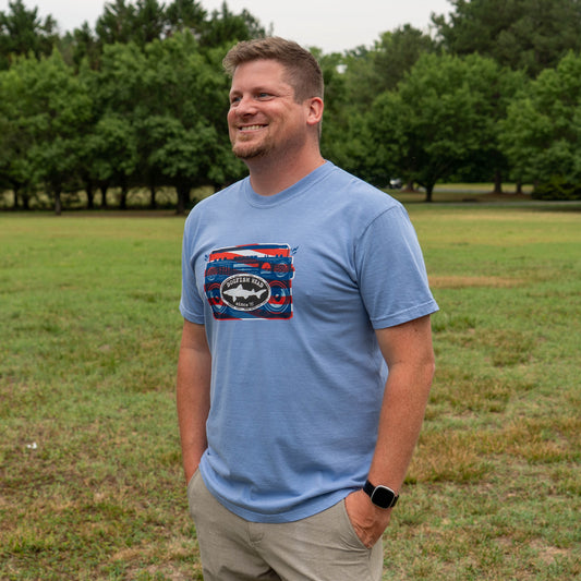 Man wearing a blue t-shirt with a graphic design standing in a grassy field.
