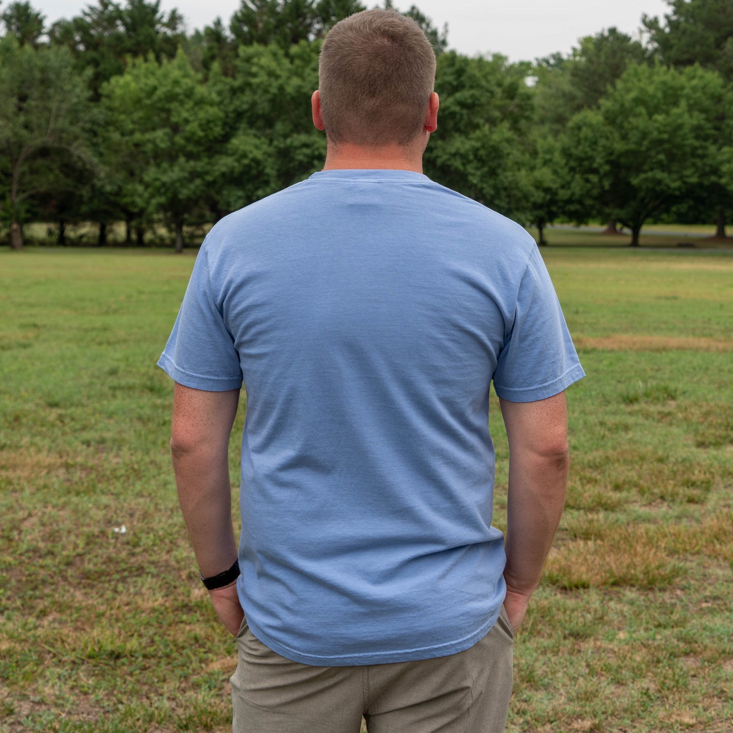 Man wearing a blue t-shirt standing in a grassy field with trees in the background