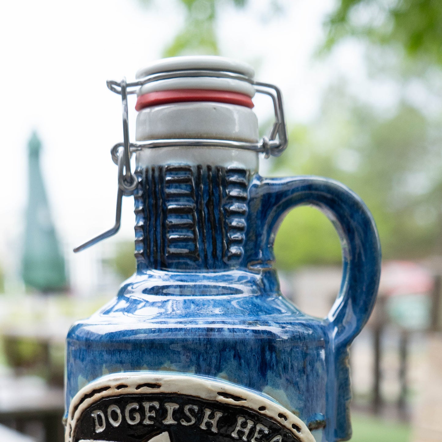 A close-up of the Blue Gradient Ceramic Growler by Romanick Pottery features a handle and swing-top cap, outdoors with blurred greenery and a patio umbrella in the background.