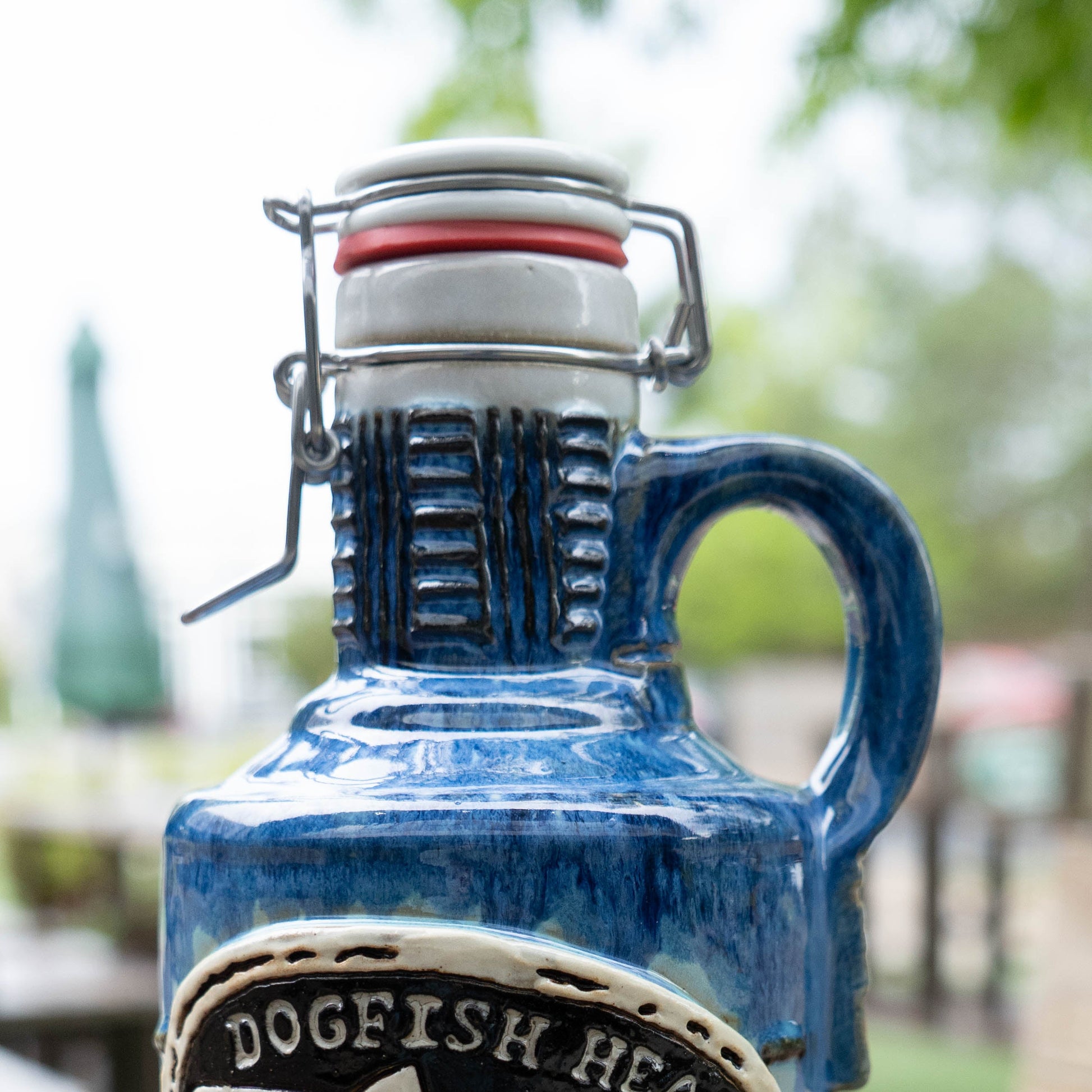 A close-up of the Blue Gradient Ceramic Growler by Romanick Pottery features a handle and swing-top cap, outdoors with blurred greenery and a patio umbrella in the background.