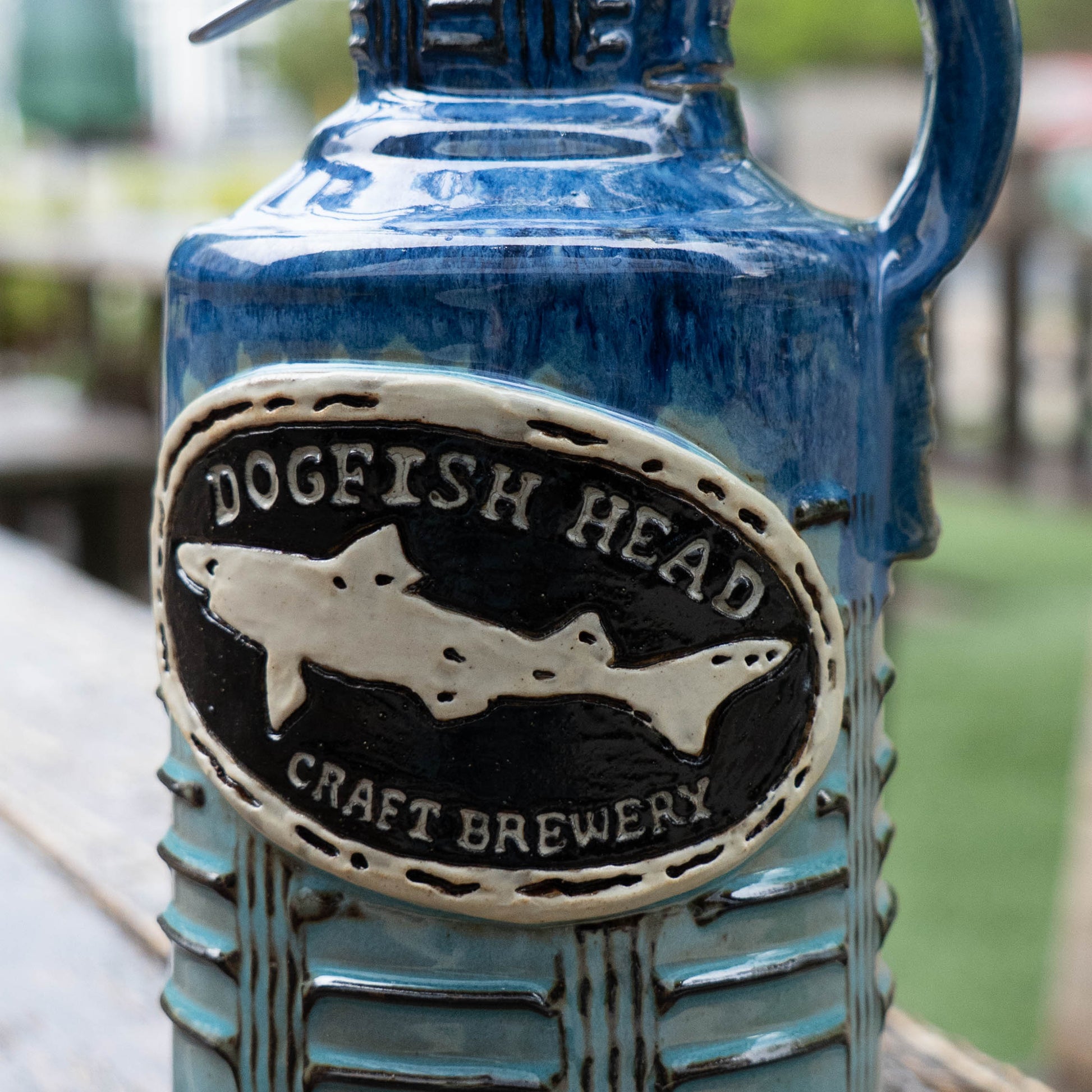 A close-up of the Blue Gradient Ceramic Growler by Romanick Pottery, featuring an oval "Dogfish Head Craft Brewery" label with a shark design. The blurred background shows outdoor seating and greenery.