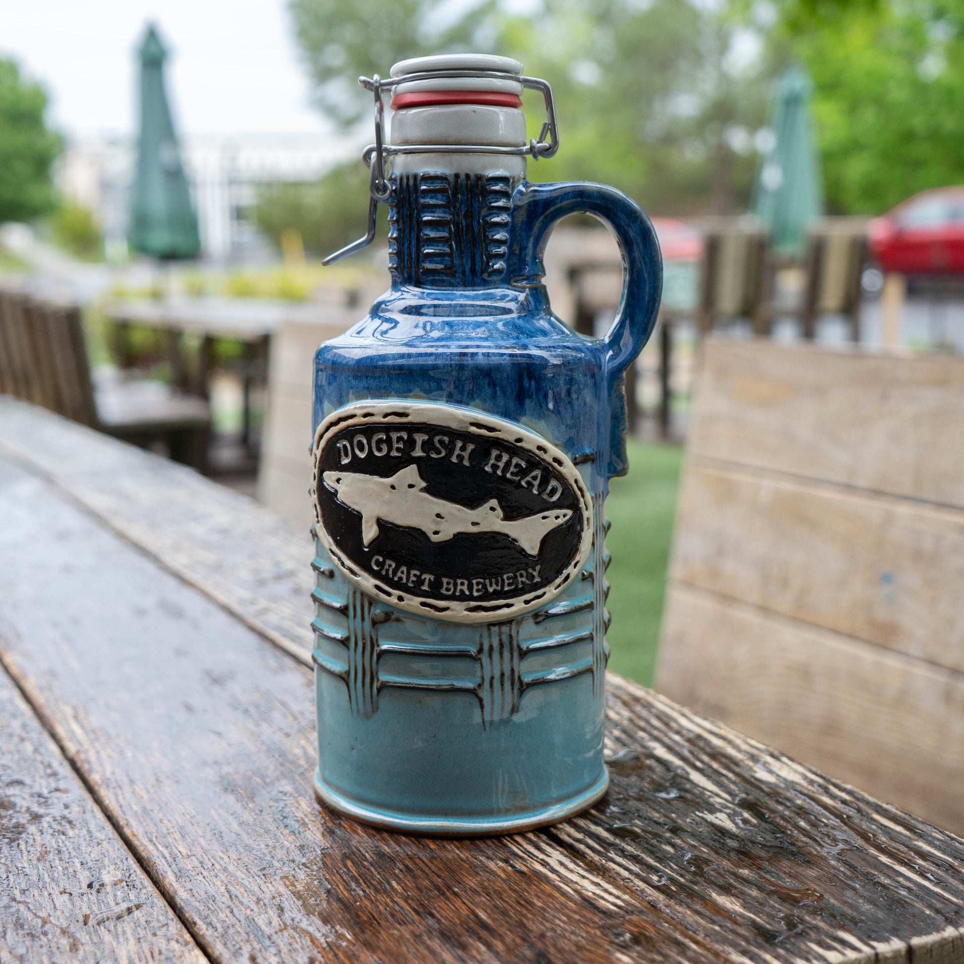 A Blue Gradient Ceramic Growler from Romanick Pottery, featuring a handle and metal clasp, rests on a weathered wooden outdoor table with blurred patio seating and greenery in the background.