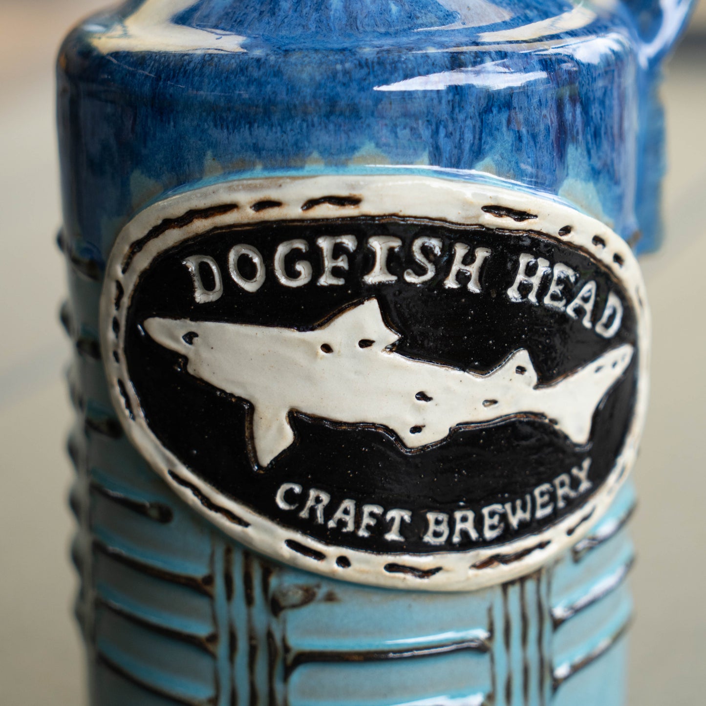 Close-up of the Blue Gradient Ceramic Growler by Romanick Pottery, featuring a raised oval Dogfish Head Craft Brewery logo and a white dogfish shark on a blue and black background.