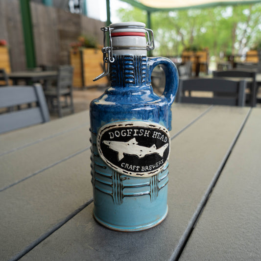 A Romanick Pottery Blue Gradient Ceramic Growler rests on a gray outdoor table; blurred chairs, tables, and greenery appear in the background.
