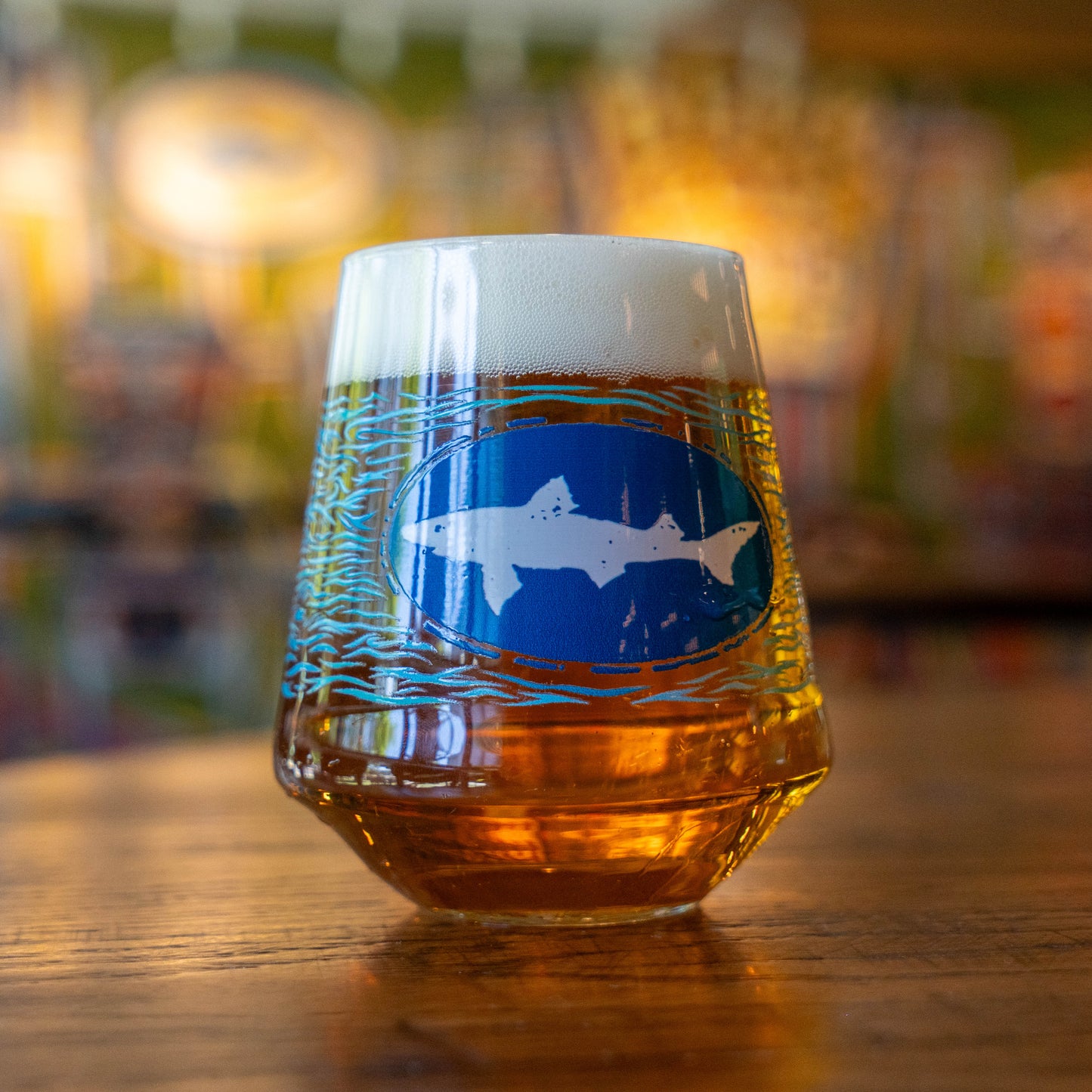 A Blue Waves Stemless Glass by Grandstand Glassware & Apparel, filled with amber craft beer and topped with foam, sits on a wooden table. The glass displays a blue logo with a white shark; the colorful background is blurred.