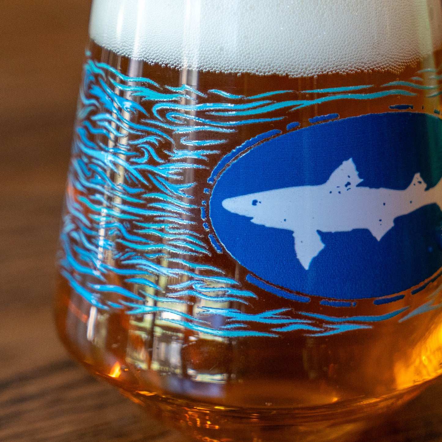 A Blue Waves Stemless Glass by Grandstand Glassware & Apparel, featuring a blue shark logo and wavy lines, holds amber craft beer with foam and rests on a wooden surface.