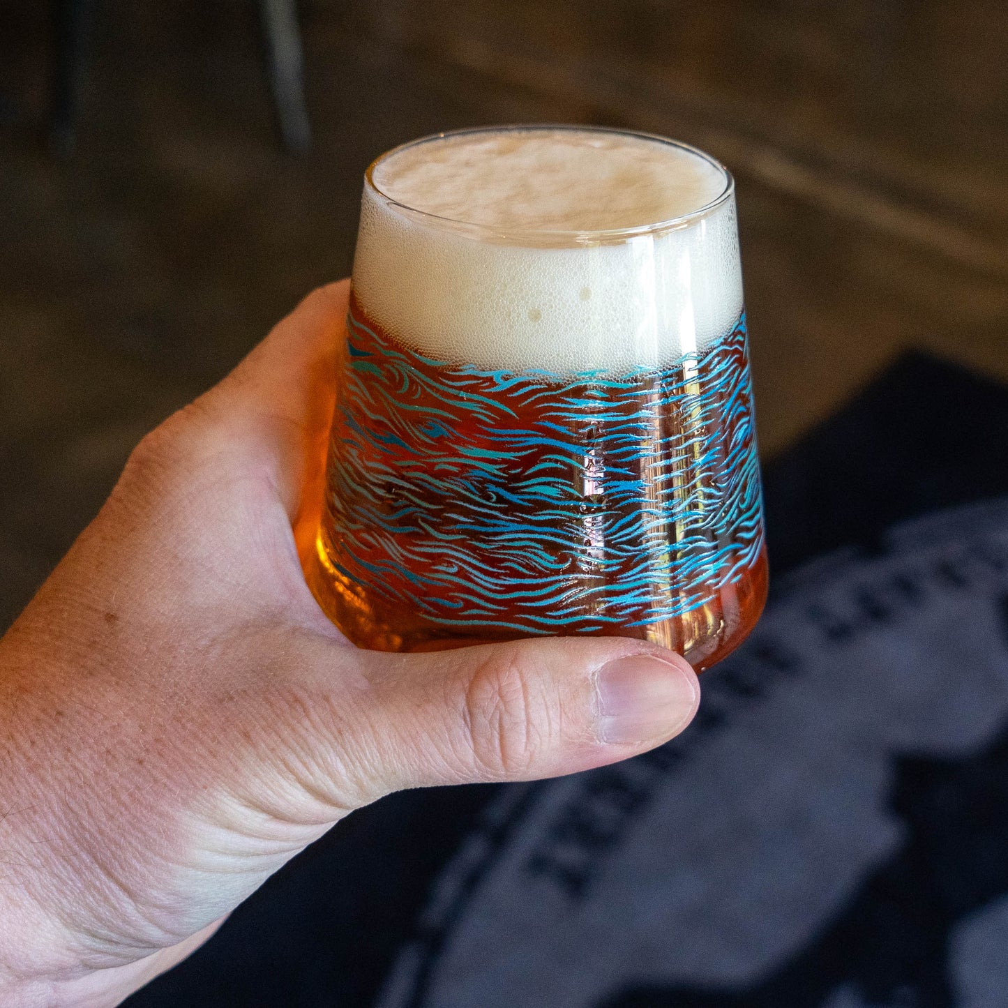 A hand holds a Blue Waves Stemless Glass from Grandstand Glassware & Apparel, filled with craft beer and topped with a thick, foamy head. The glass features a vibrant blue and orange wavy pattern; the blurred background shows part of the Dogfish Head logo.