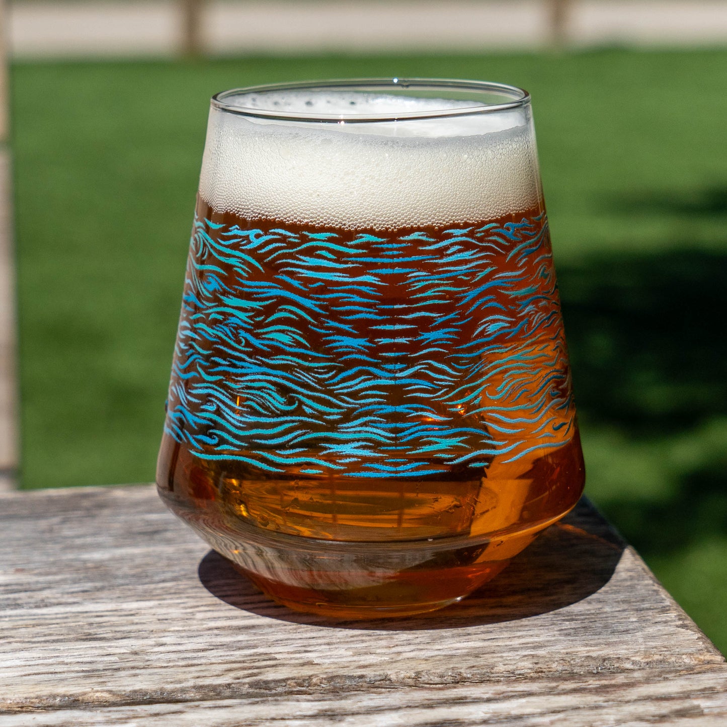 A Blue Waves Stemless Glass by Grandstand Glassware & Apparel, filled with amber craft beer and a foamy head, rests on a wooden surface. The artistic blue wave design contrasts with the grassy, sunlit outdoors for an inviting scene.
