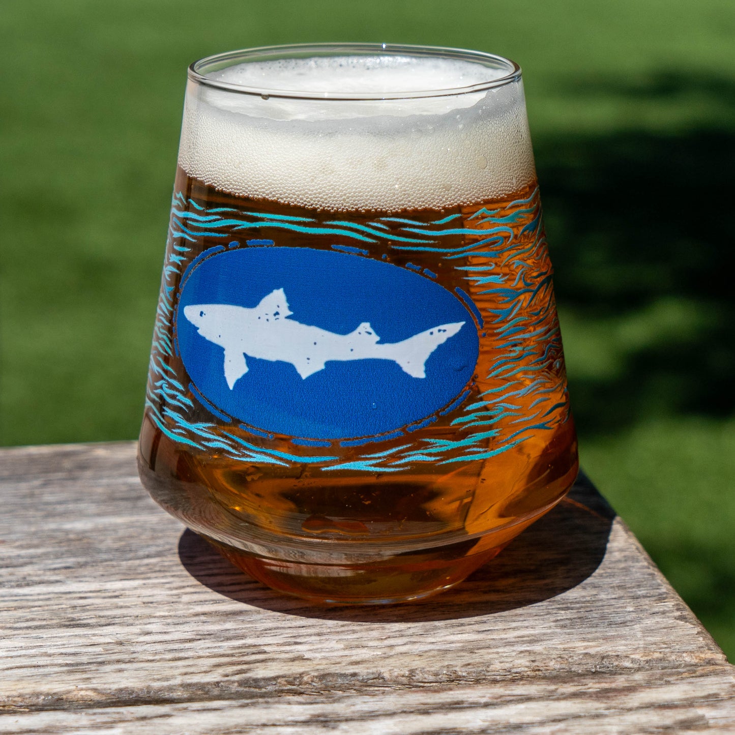 A glass of amber craft lager with a foamy head sits on a wooden surface outdoors. The Blue Waves Stemless Glass by Grandstand Glassware & Apparel, decorated with blue, orange, and teal waves, is featured. Green grass fills the background.