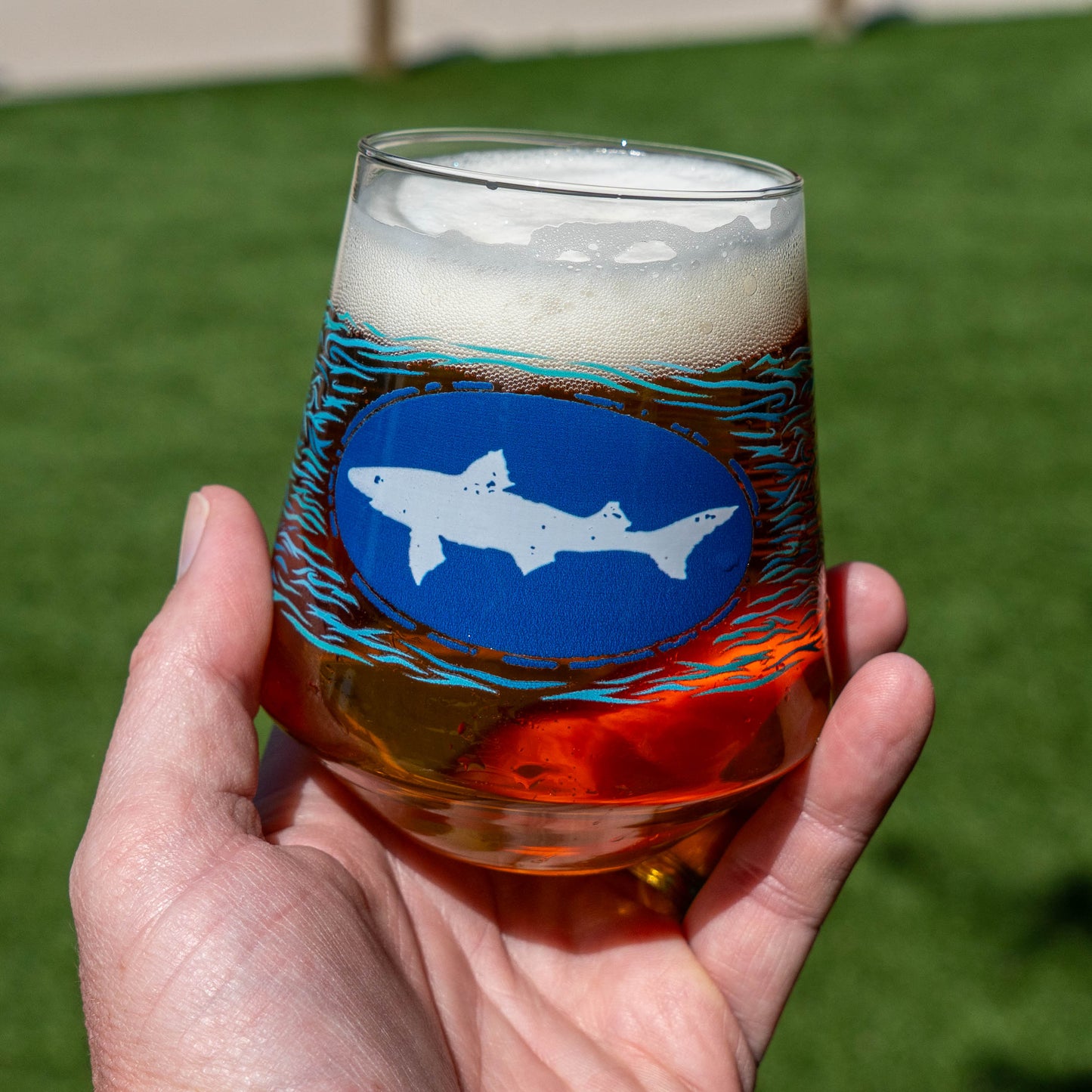 A hand holds a Blue Waves Stemless Glass from Grandstand Glassware & Apparel, filled with amber craft lager and topped with foam. The glass shows a blue oval logo, while a blurred green lawn appears in the background.