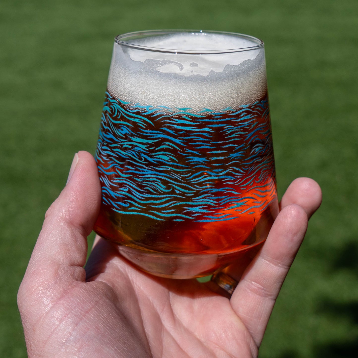 A hand holds Grandstand Glassware & Apparel’s Blue Waves Stemless Glass, filled with amber craft beer and topped with foam, set against a blurred green grassy background. The glass features a distinctive blue wavy pattern.