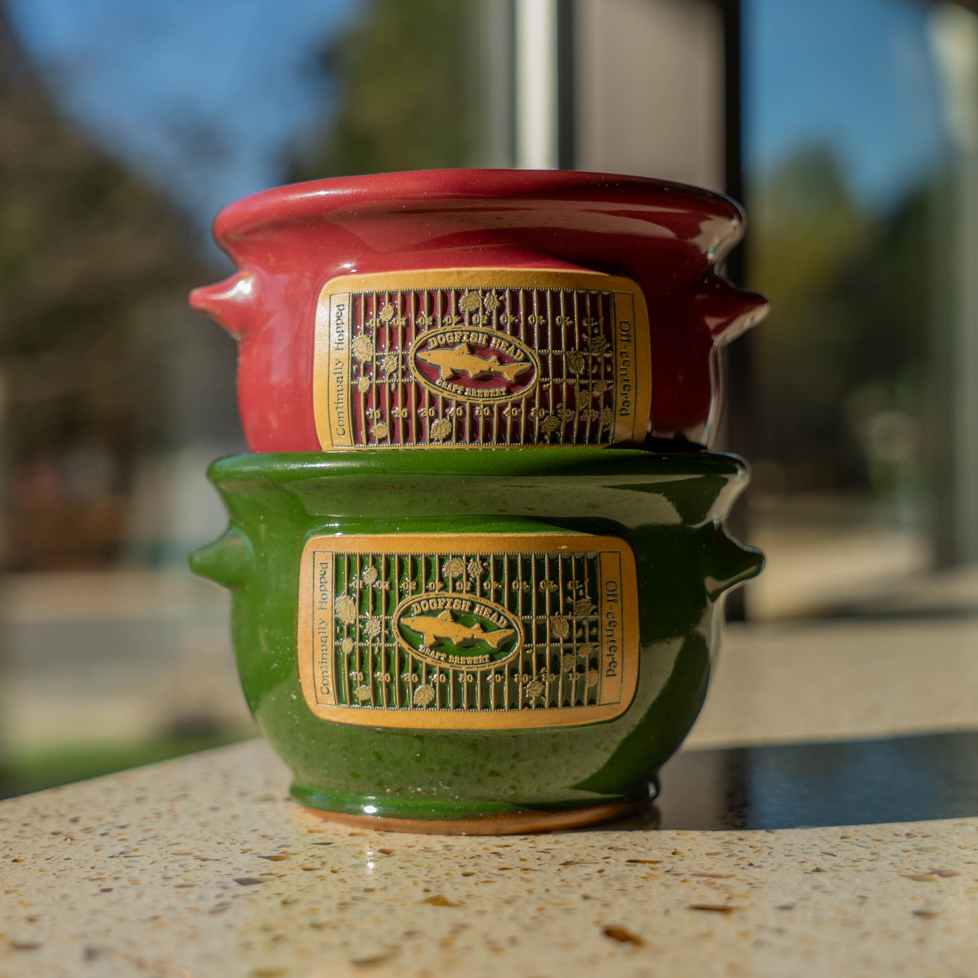 Stack of red and green ceramic crocks with branding on a blurred outdoor background