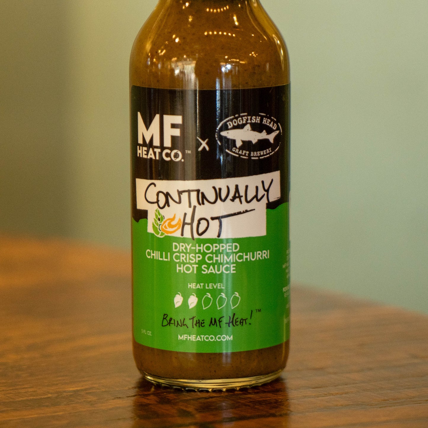 A bottle of Continually Hot Sauce by MF Heat Company is on a wooden surface, highlighting its moderate heat and vibrant chimichurri flavors from the Dogfish Head collaboration.