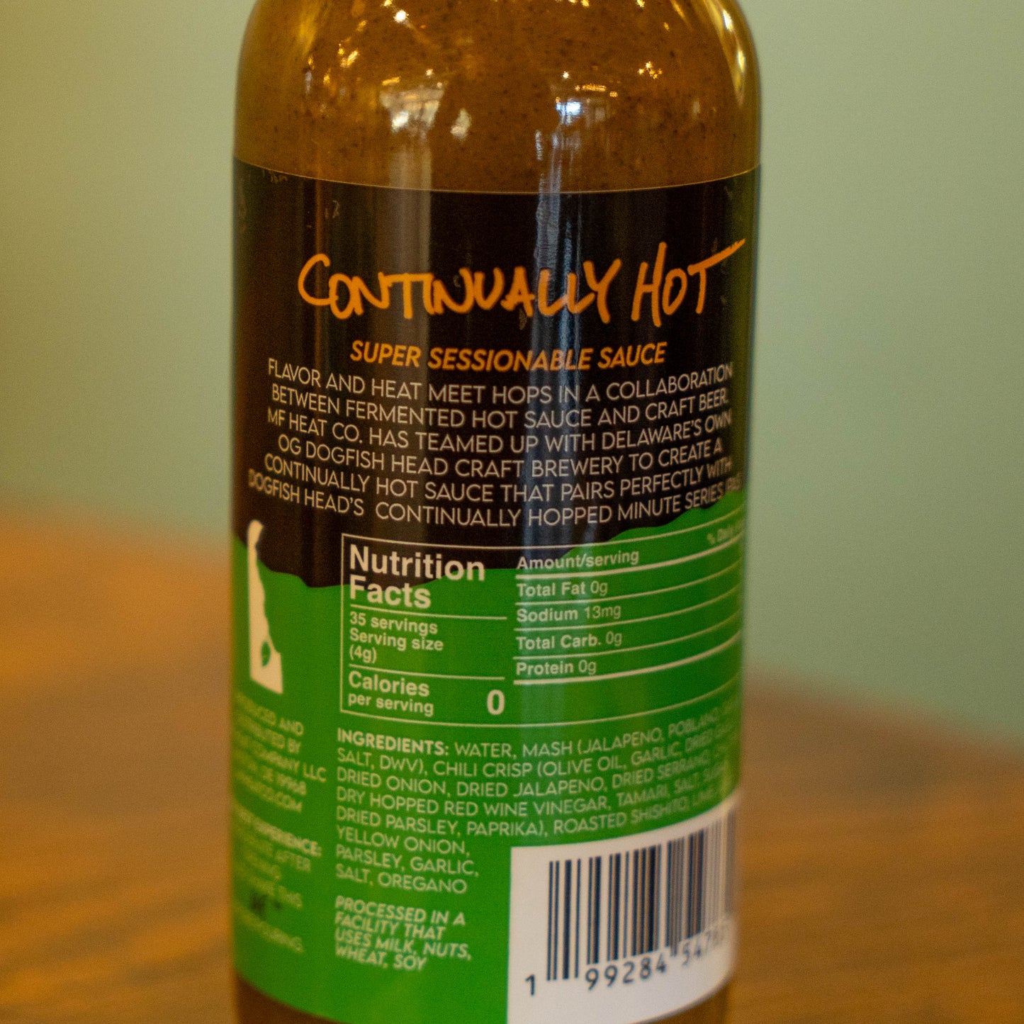 A close-up of a bottle labeled Continually Hot Sauce from MF Heat Company, featuring nutrition facts and ingredients on a green and black background, sitting on a wooden surface.