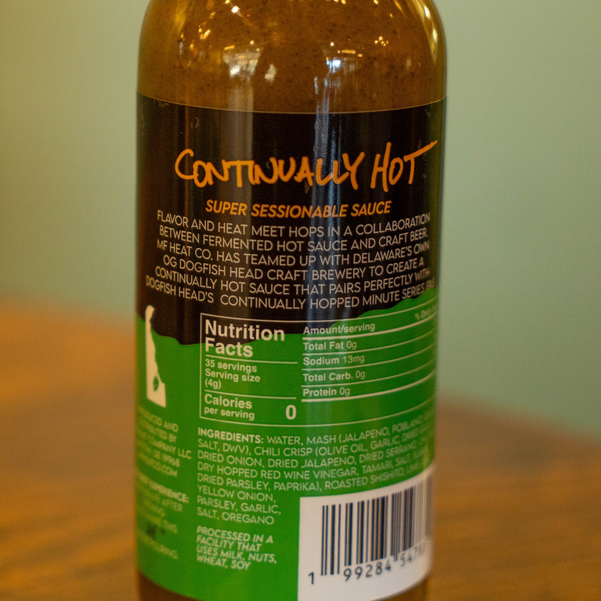 A close-up of a bottle labeled Continually Hot Sauce from MF Heat Company, featuring nutrition facts and ingredients on a green and black background, sitting on a wooden surface.