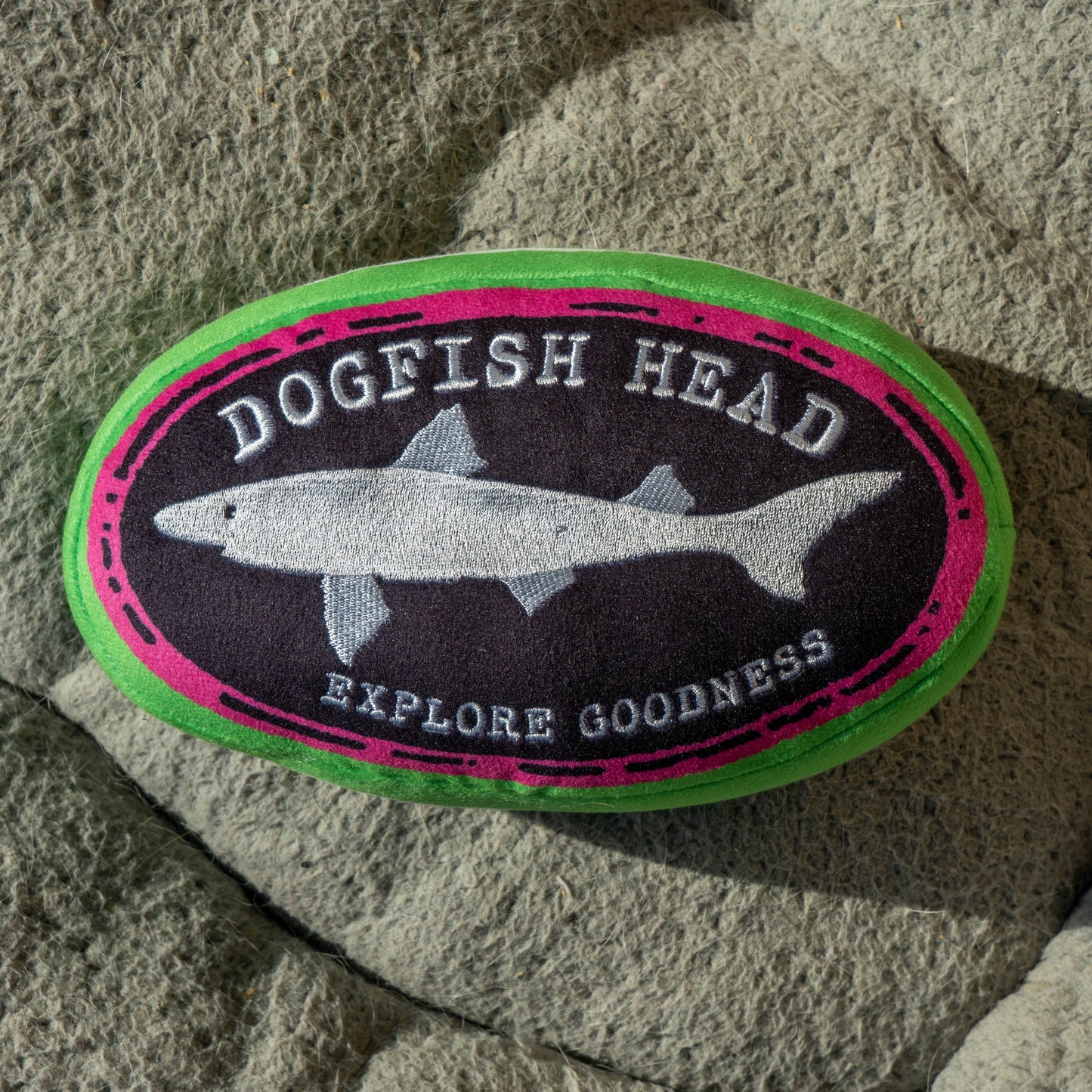 Oval shaped Dogfish Head plush dog toy with a black logo that reads "Dogfish Head, Explore Goodness". The logo is outlined in hot pink and then outlined and covered in green.