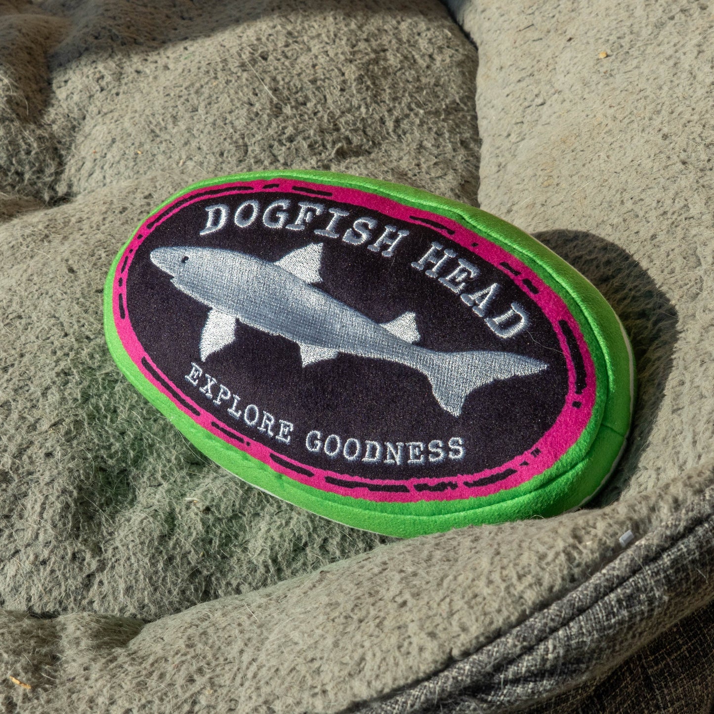 Oval shaped Dogfish Head plush dog toy with a black logo that reads "Dogfish Head, Explore Goodness". The logo is outlined in hot pink and then outlined and covered in green.