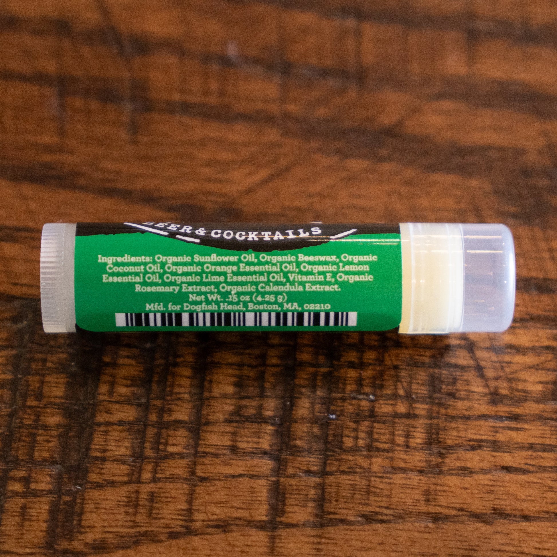 A tube of Eco Lips Inc Lip Balm labeled Beer & Cocktails, with a green label listing organic oils and extracts, rests on a wooden surface. Product information and the company address are also visible.