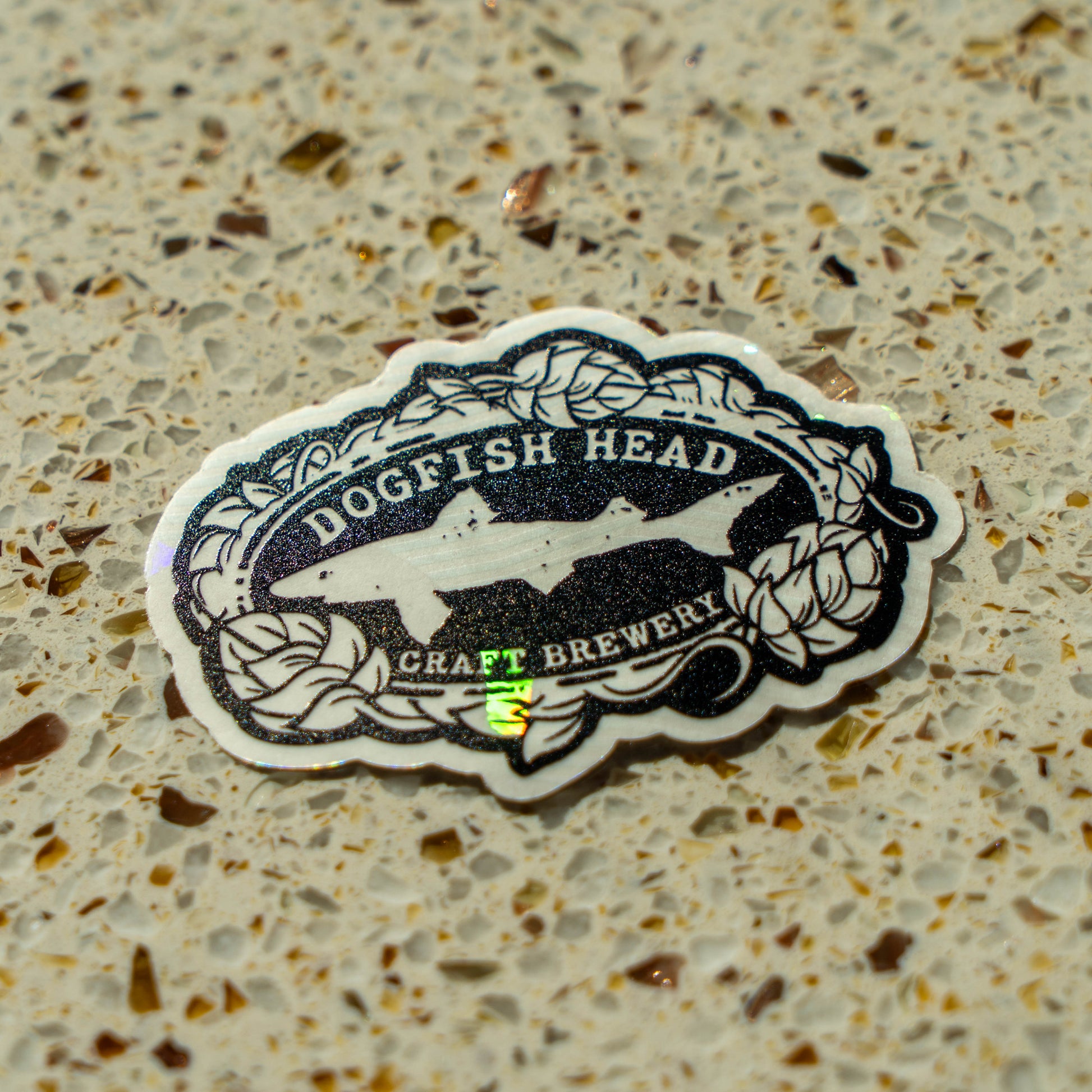 A black and white Rainbow Symphony Suncatcher Logo Sticker with a shark logo and leaf border, perfect for brewery gear fans, sits on a speckled beige countertop.