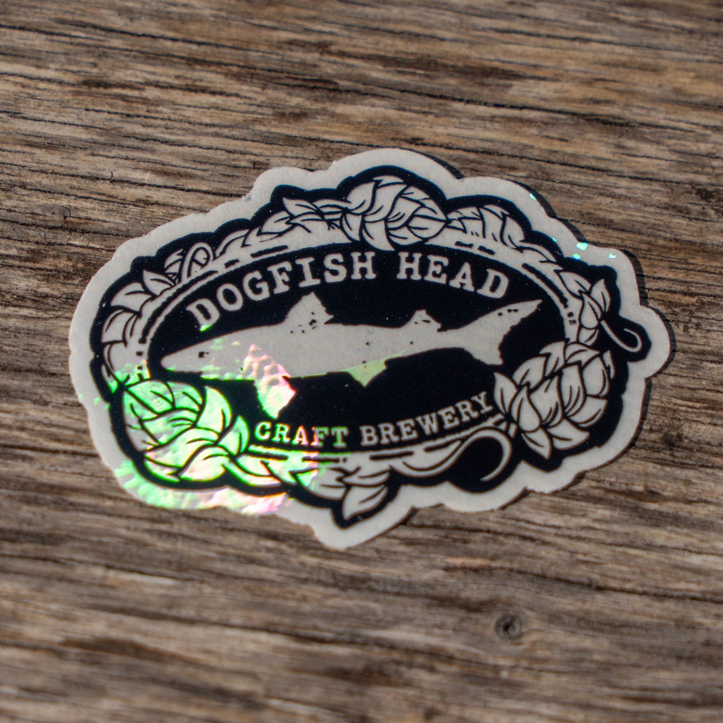 The Suncatcher Logo Sticker by Rainbow Symphony features the Dogfish Head logo with a fish in an oval surrounded by hop vines, displayed on textured wood—an ideal addition to your brewery gear collection.