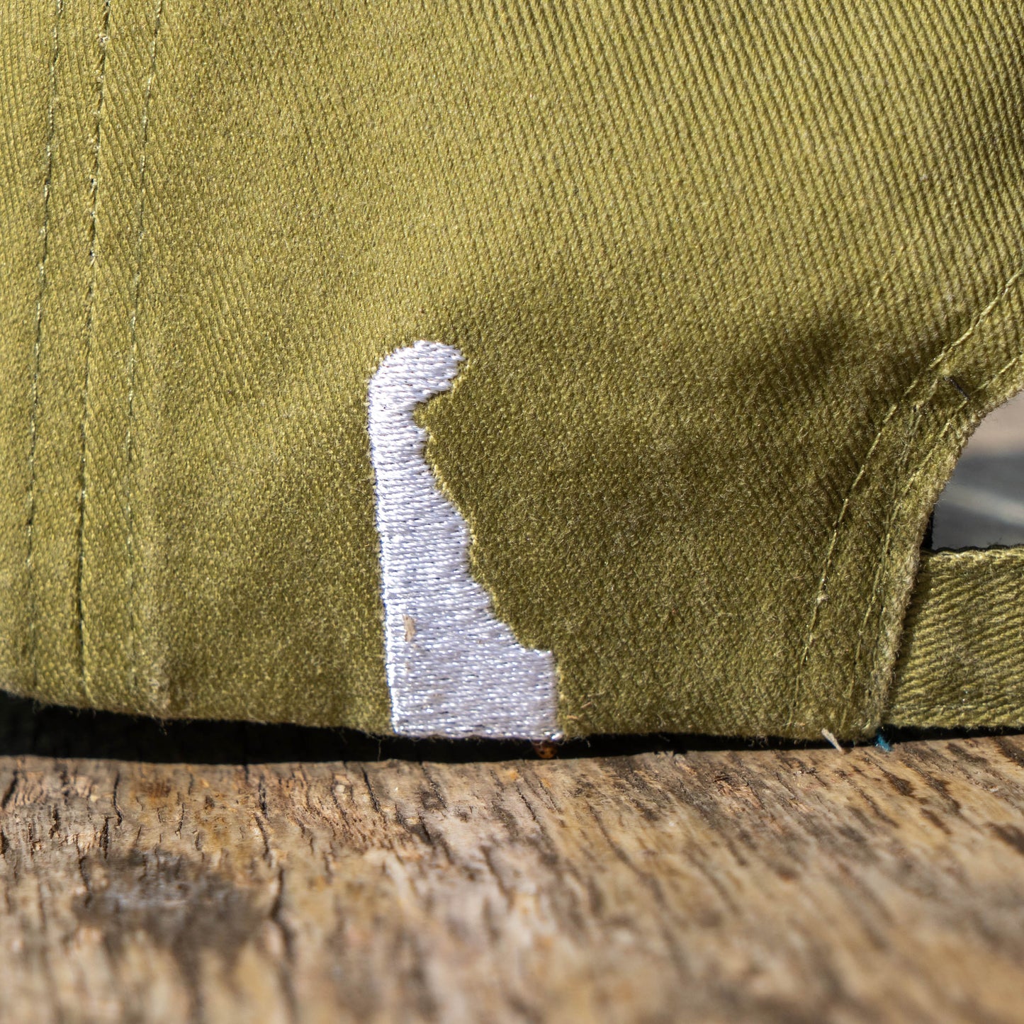 Close up of white embroidered shape that represents that state of Delaware on an olive green dad hat