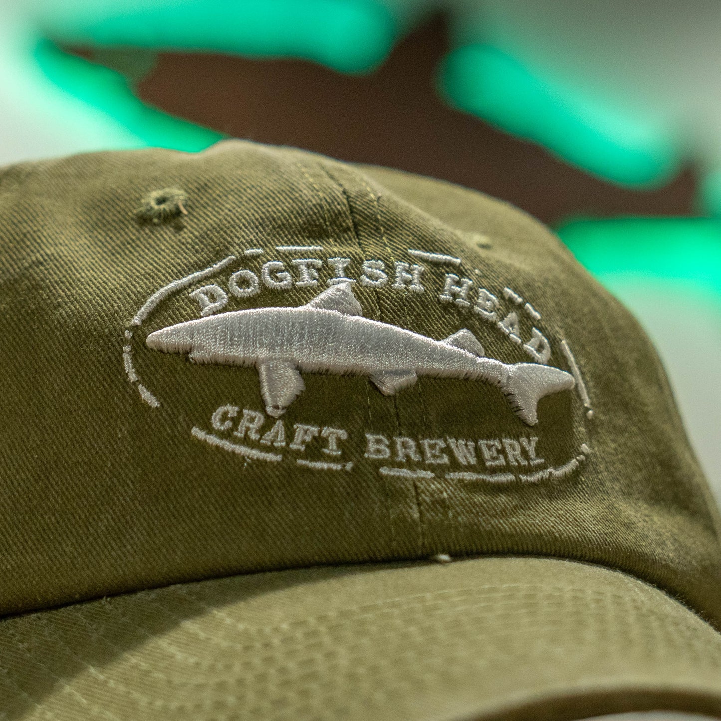 Close up of white Dogfish Head Craft Brewery embroidered logo on an olive colored dad hat 